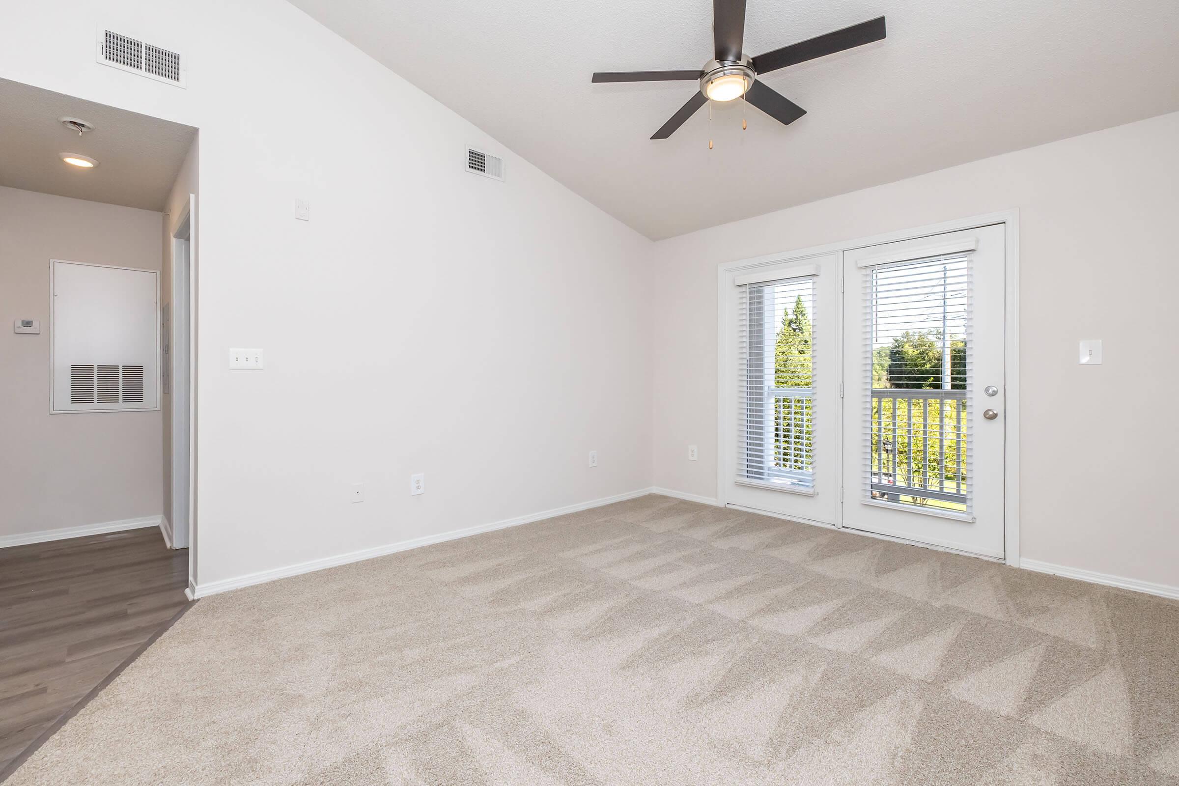 Spacious, well-lit living area with vaulted ceiling, ceiling fan, and large windows. Light-colored walls and soft carpet create a warm atmosphere. Two sliding glass doors lead to a balcony with views of greenery outside. Modern design with neutral tones, offering a comfortable and inviting space.