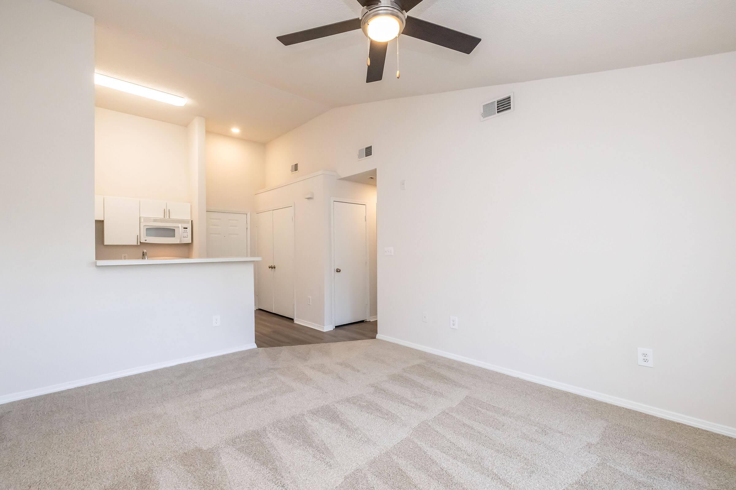 A spacious, well-lit living area featuring light-colored walls and a ceiling fan. The room has a plush carpet and an open layout that connects to a kitchen space with modern appliances. There are two doors leading to additional rooms, and ample natural light comes from the windows.