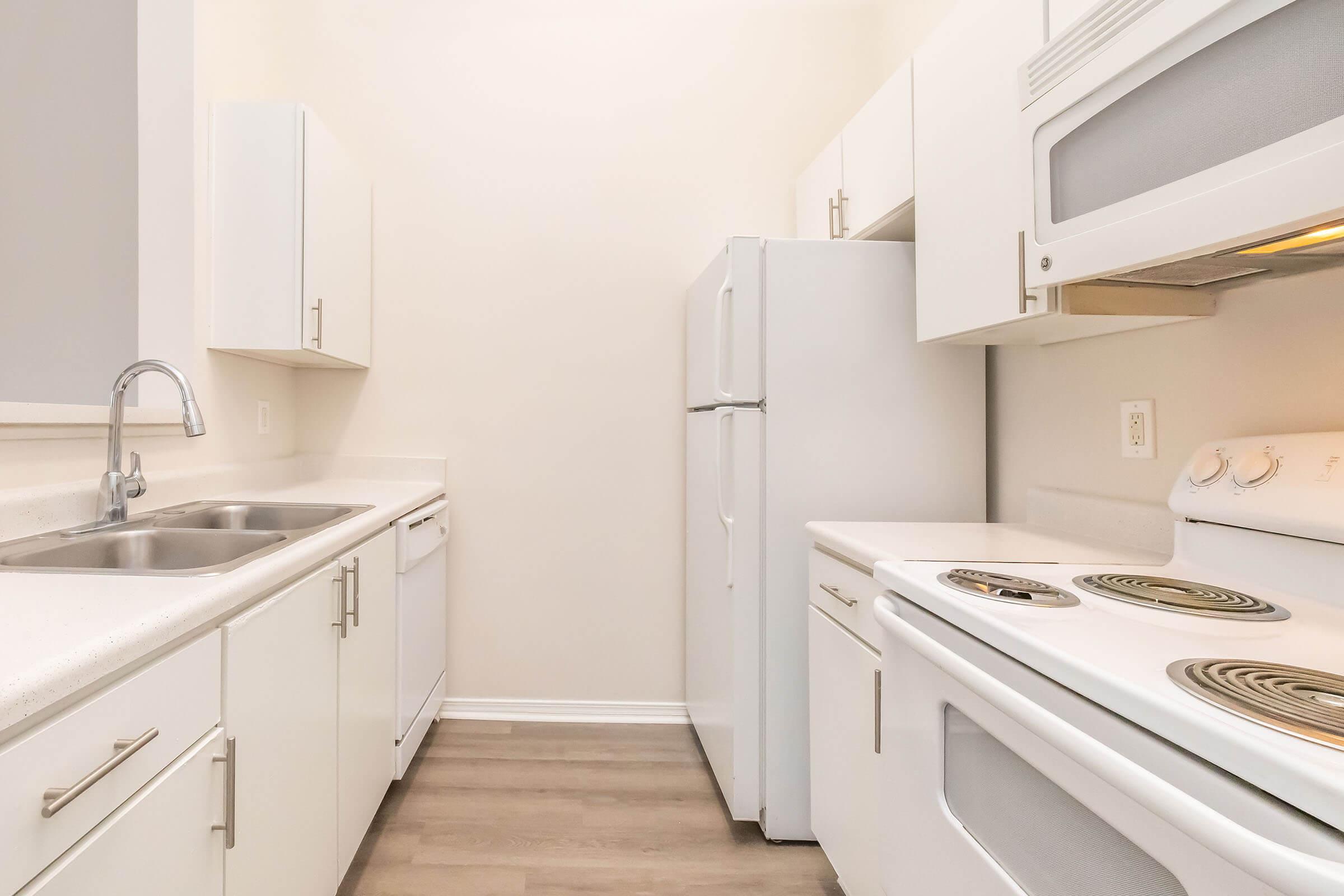 A clean and modern kitchen featuring white cabinetry, a double sink, a dishwasher, a refrigerator, and a stove with an oven. The walls are light-colored, and the flooring is a light laminate, creating a bright and spacious atmosphere.