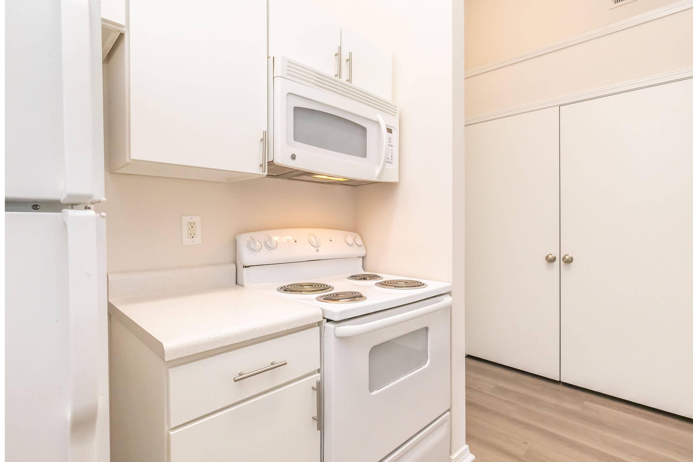 A small, modern kitchen featuring a white stove and microwave set against light-colored cabinets. The countertop is clean and minimalistic, with a refrigerator on the left and a closed closet door on the right, creating a tidy and functional space.