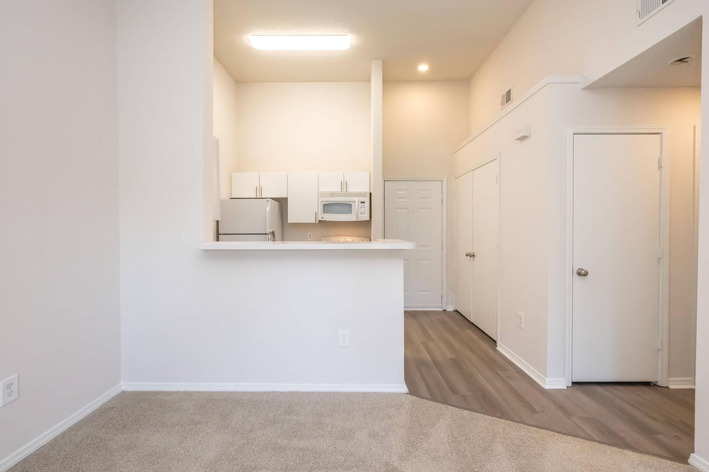 A bright, modern apartment interior featuring a carpeted living area with a small kitchen area visible in the background. The kitchen has white cabinets, an oven, a refrigerator, and a microwave. A closed door leads to another room, with neutral-colored walls and ample natural light.