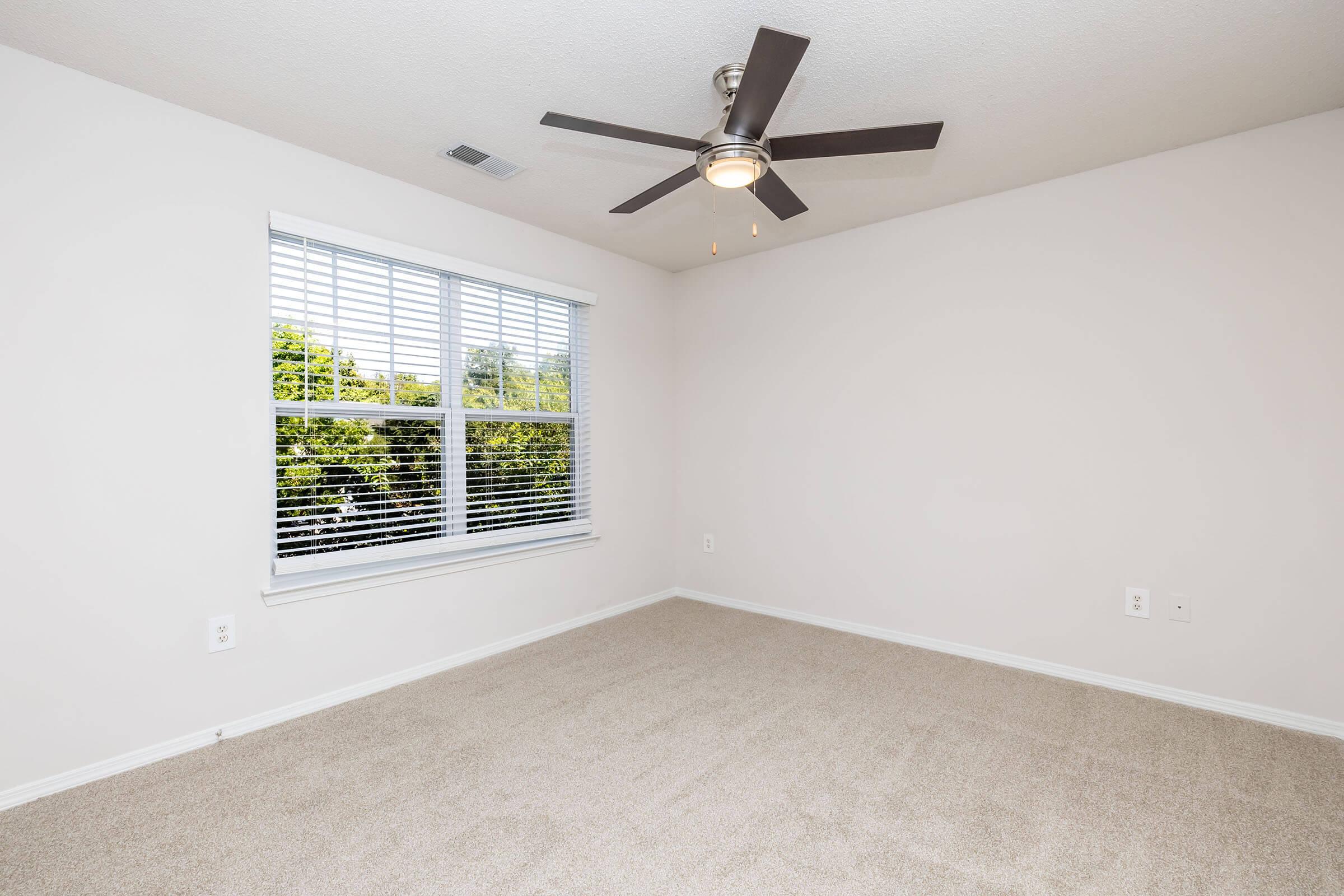 Empty room with light beige carpet and white walls. A ceiling fan with lights is installed in the center. There is a window with white blinds that lets in natural light, showing greenery outside. The space is clean and minimalistic, suitable for various furniture arrangements.