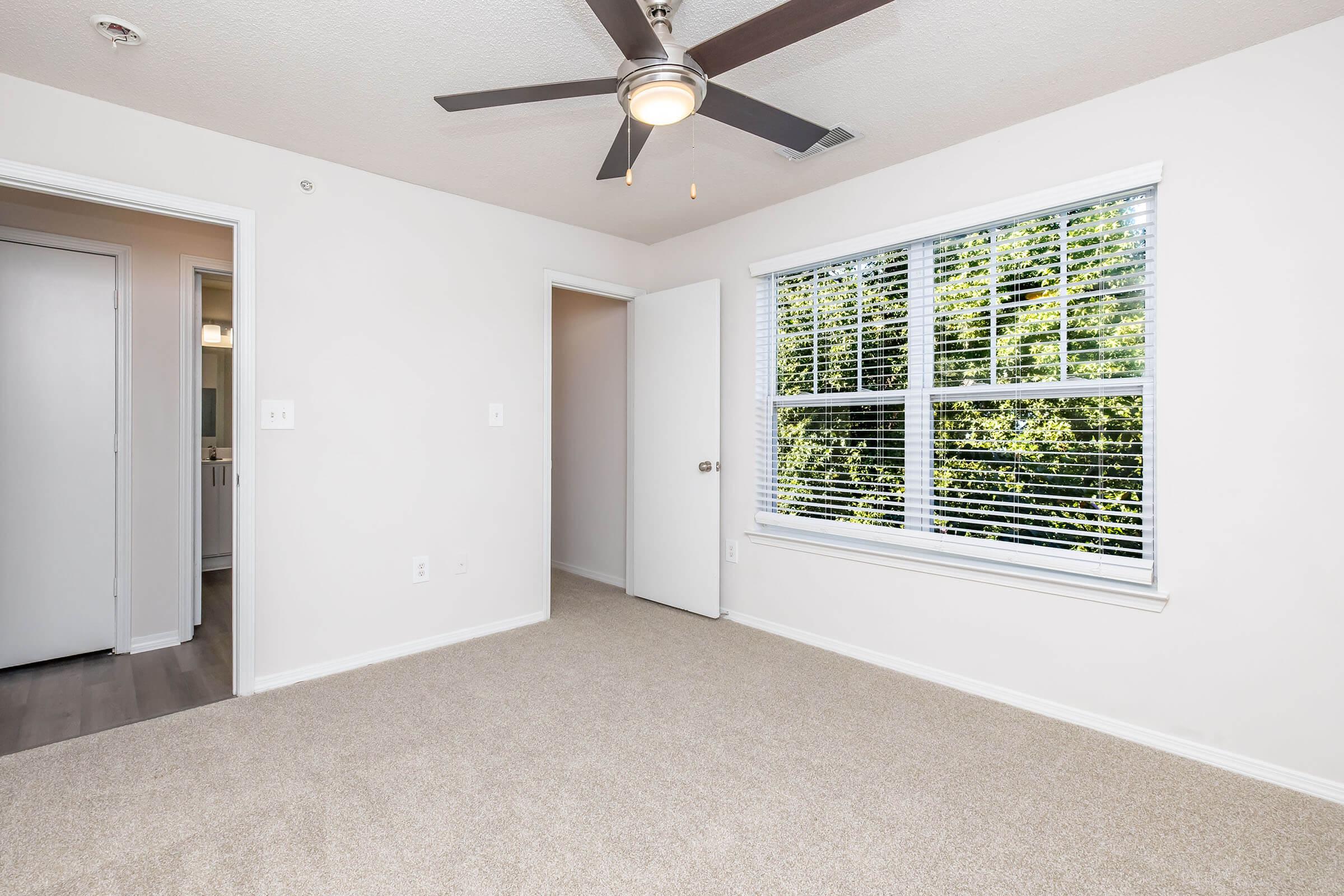 A well-lit, empty bedroom featuring a ceiling fan, a large window with blinds, and beige carpeting. The room has white walls and two doorways leading to adjacent spaces. Natural light shines through the window, creating a spacious and inviting atmosphere.