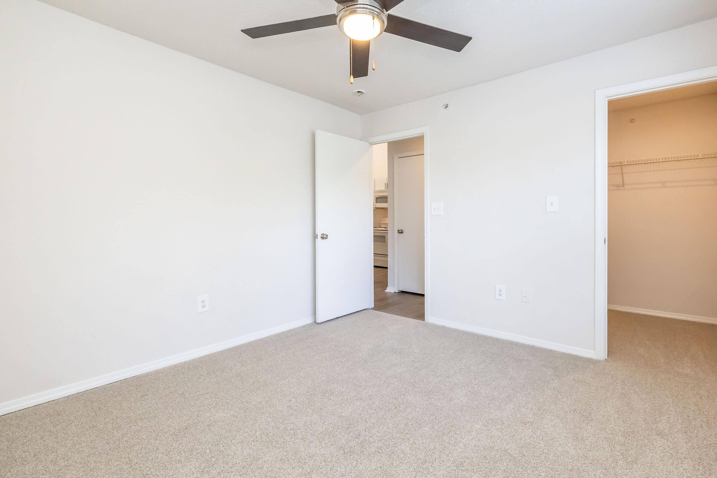 A bright, empty room with beige carpet and white walls. There is a ceiling fan with three blades, a closed door on the left leading to another room, and an open door on the right leading to a walk-in closet. The room appears spacious and well-lit, designed for potential furnishings.