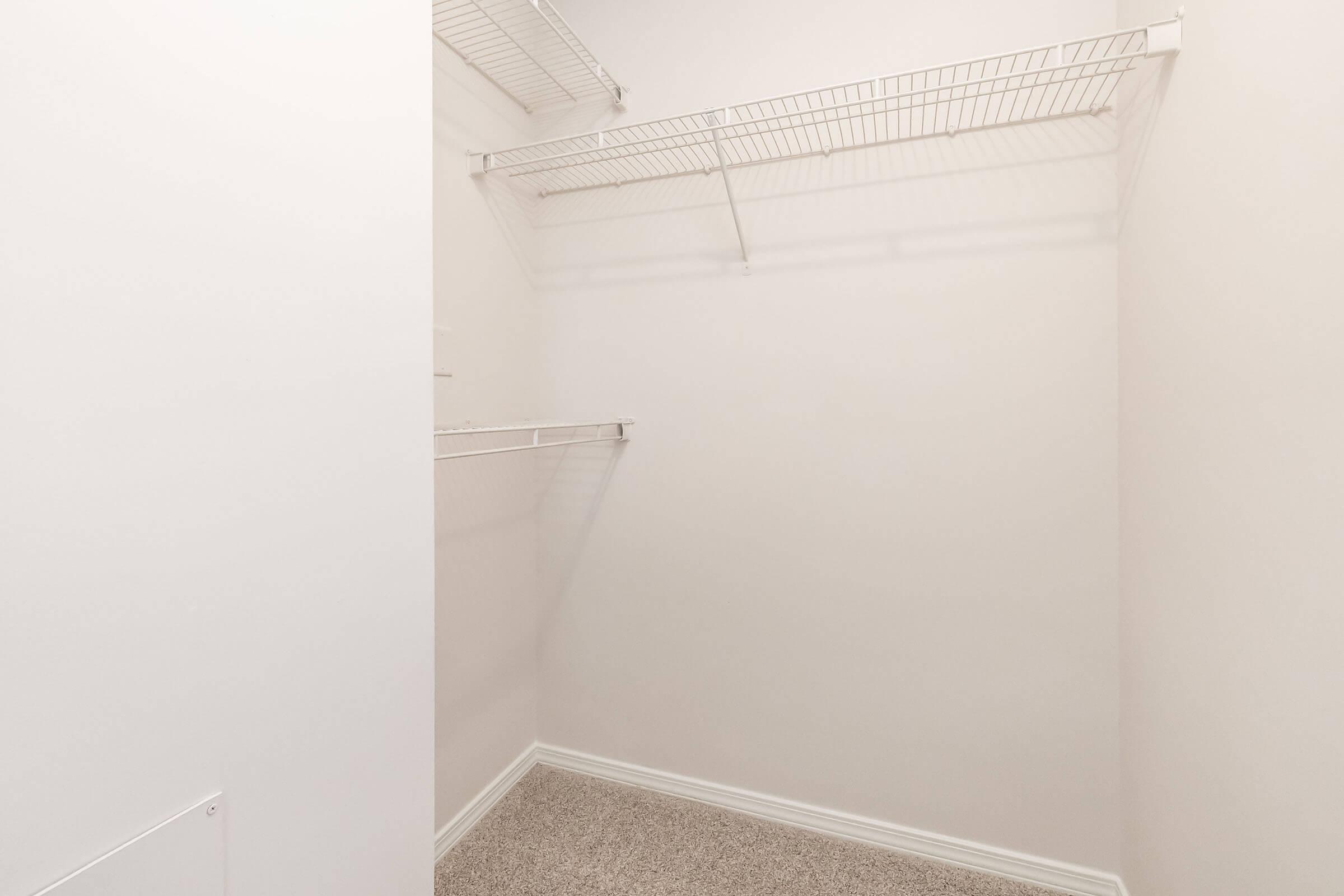 An empty closet with white walls and wire shelves. The closet features a clean, minimalist design with gray carpeting on the floor and sparse shelving, giving it a spacious appearance.