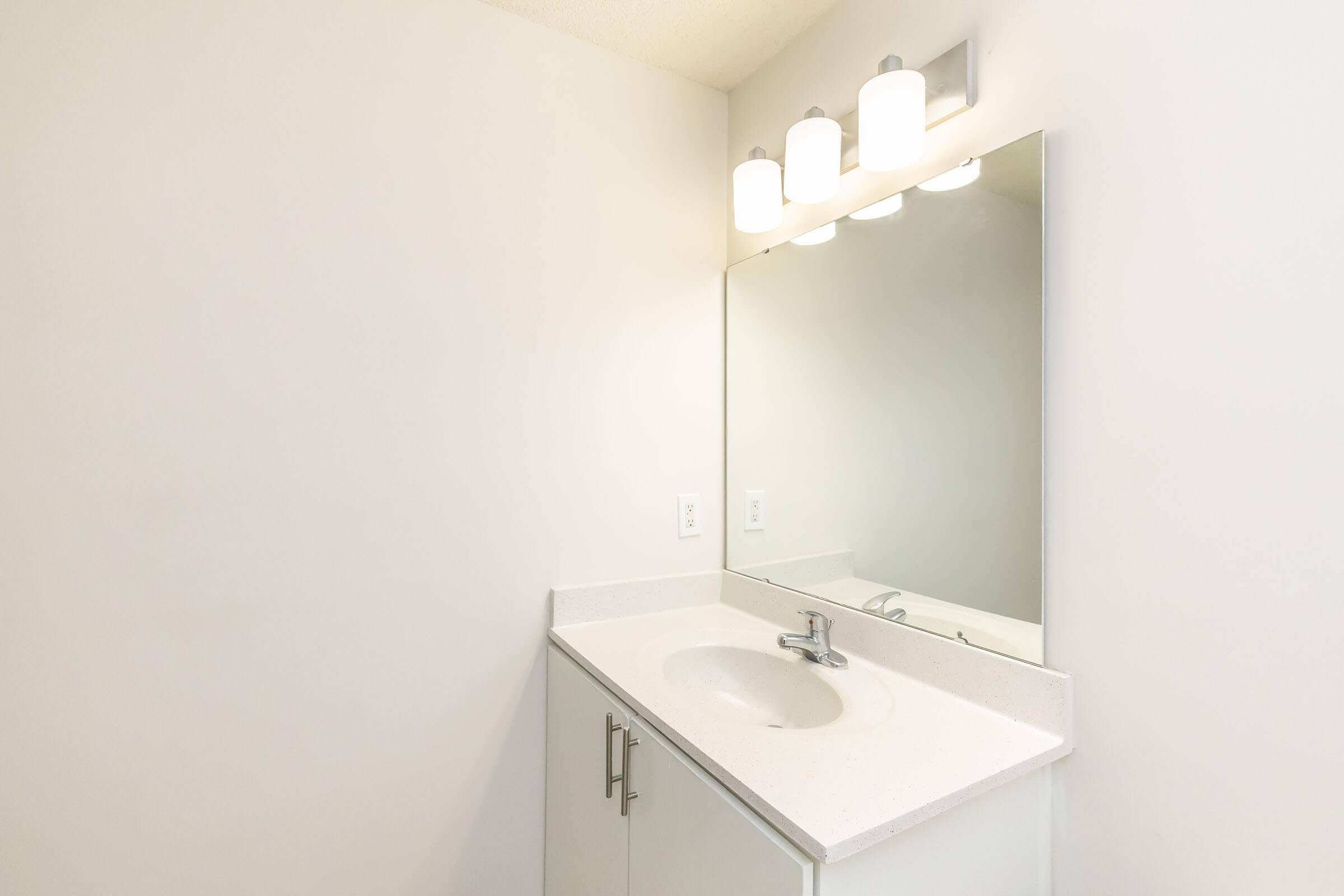 A clean, modern bathroom vanity with a white countertop and a single sink. Above the sink, there are three light fixtures providing bright illumination. The walls are painted white, and there is a large mirror reflecting the space. The overall design is minimalist and functional.