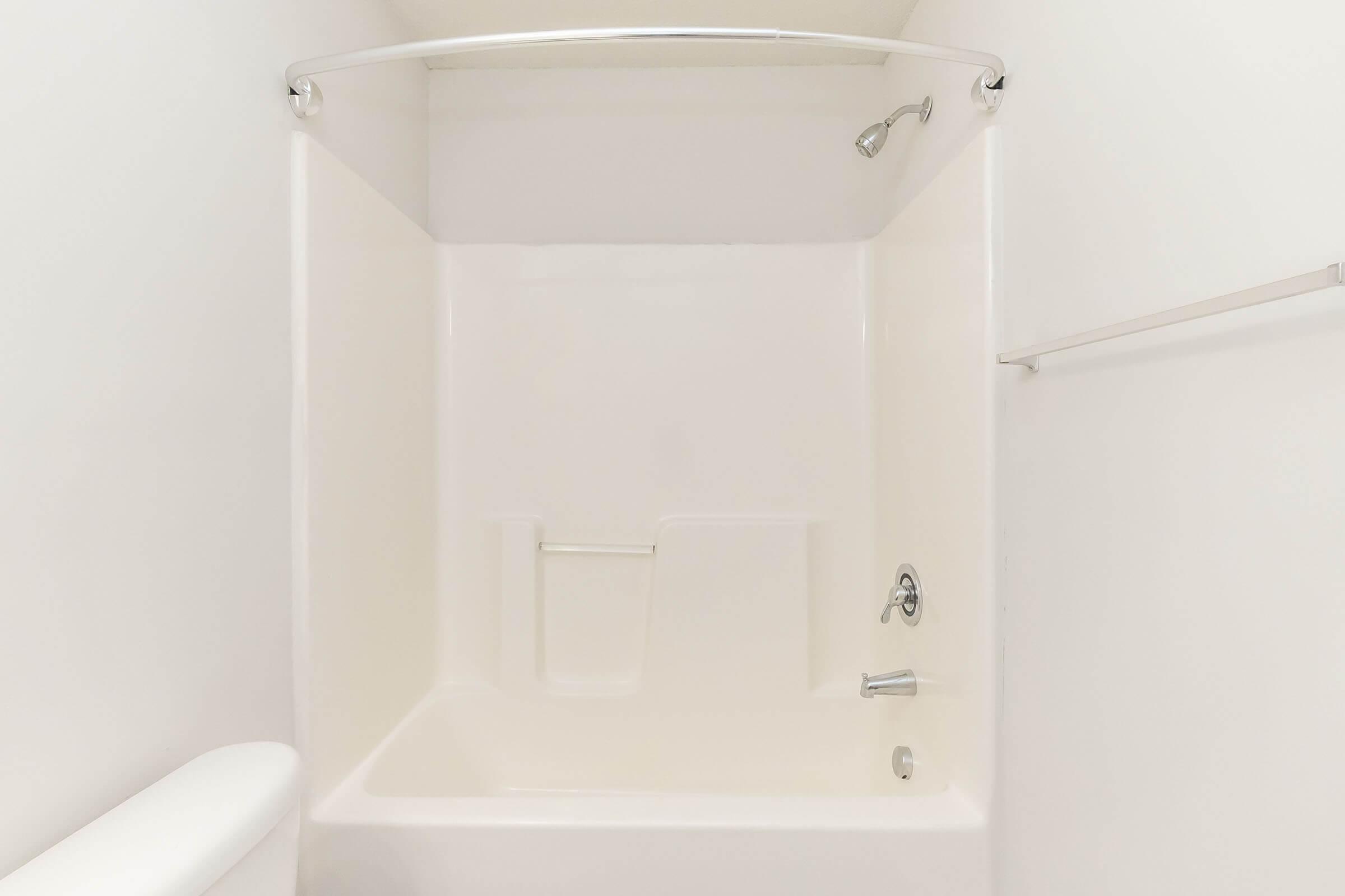 A clean, empty bathroom shower area with a white tiled wall, a curved shower rod, a metal shower head, and a single faucet. The space has minimal decor, featuring a white tub and a small shelf on the wall. Natural light illuminates the area.
