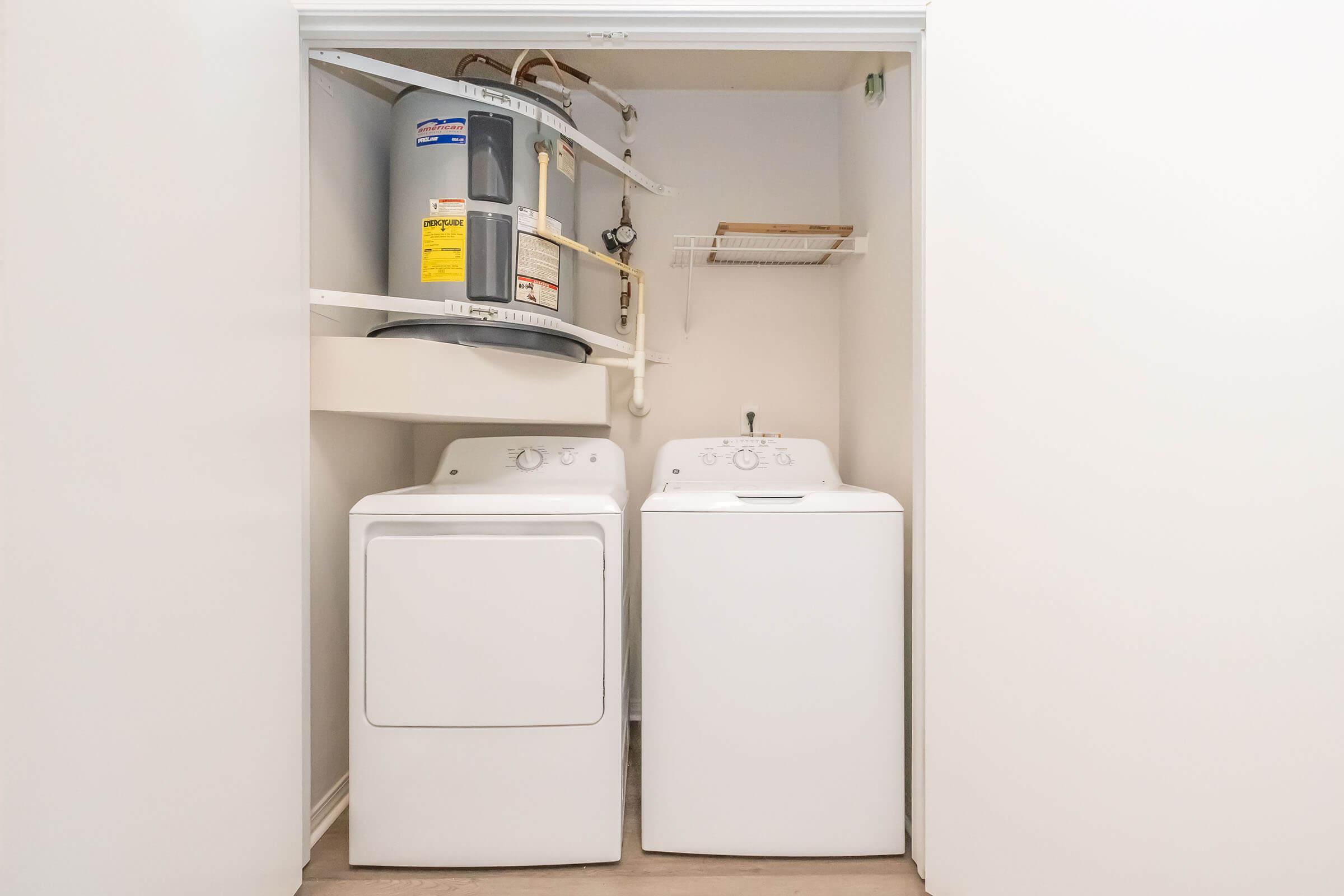 A small laundry space featuring a white washing machine and dryer side by side. Above them is a water heater mounted on the wall, along with a shelf for storage. The walls are painted white, and the floor is a light-colored laminate.