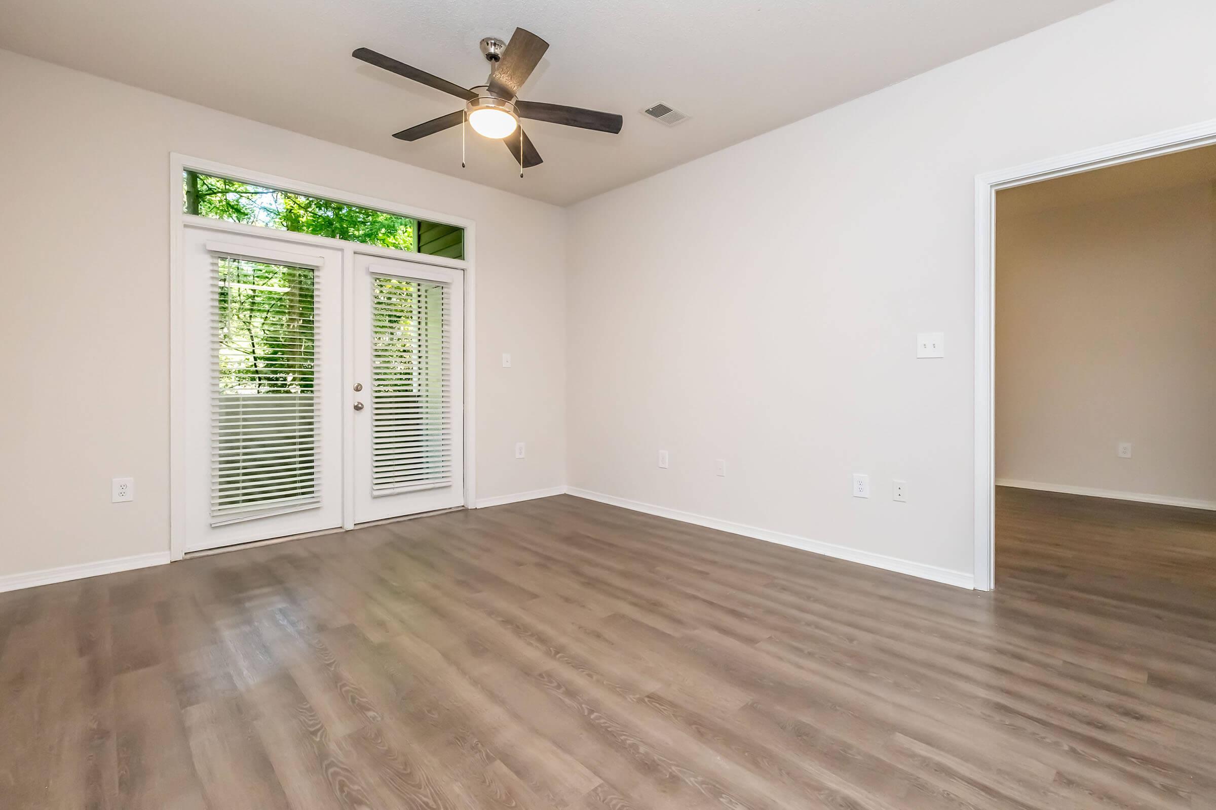 A spacious, empty living room featuring light-colored walls and medium wood flooring. A ceiling fan is mounted above, and there's a set of double doors with blinds leading to an outdoor area. To the right, an open doorway leads to another room. The overall atmosphere is bright and airy.