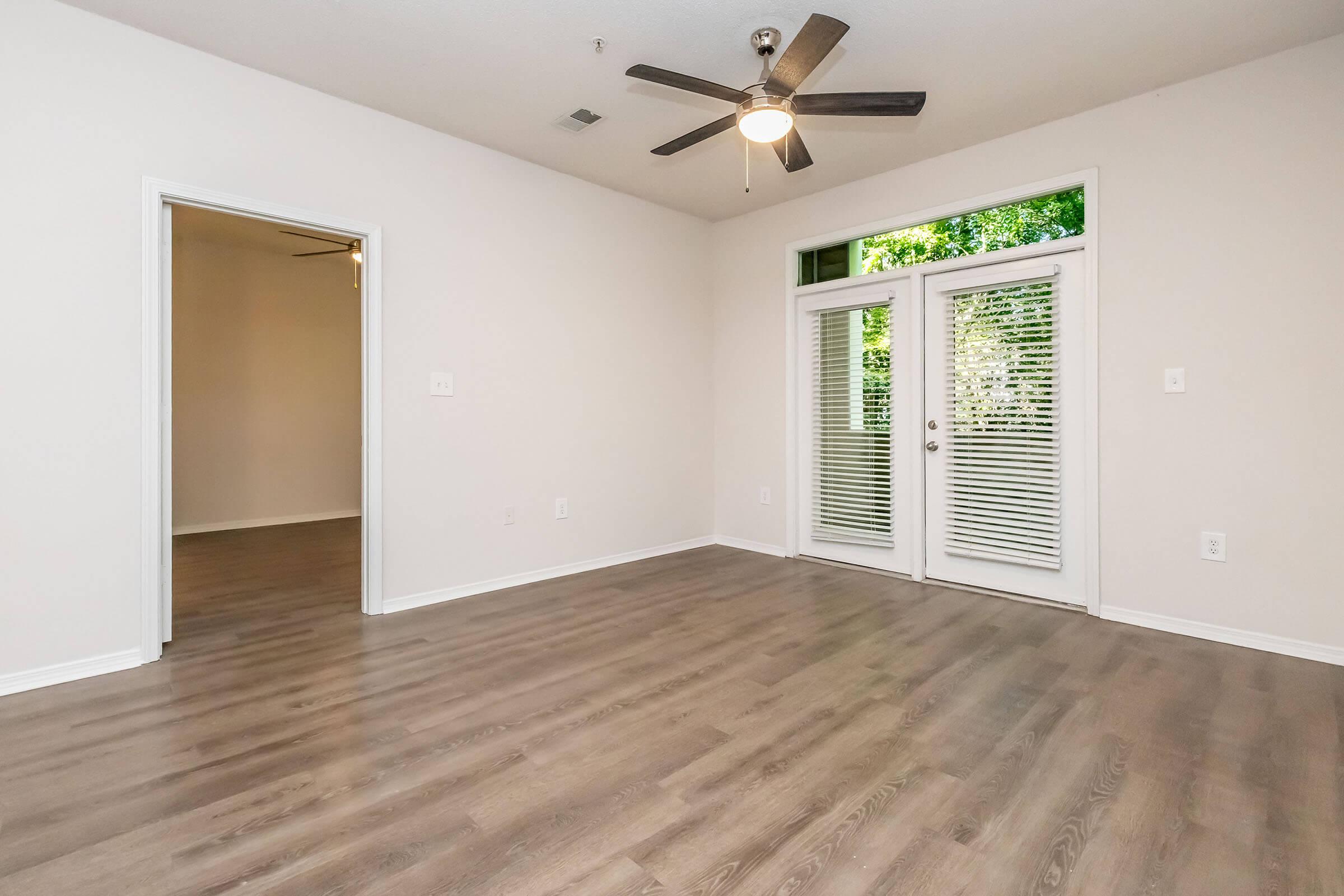 A spacious, well-lit room featuring light hardwood floors, a ceiling fan, and large windows with blinds. There are two sets of doors leading to an outdoor area. The walls are painted in a neutral color, creating a bright and inviting atmosphere. An open doorway leads to an adjacent room.