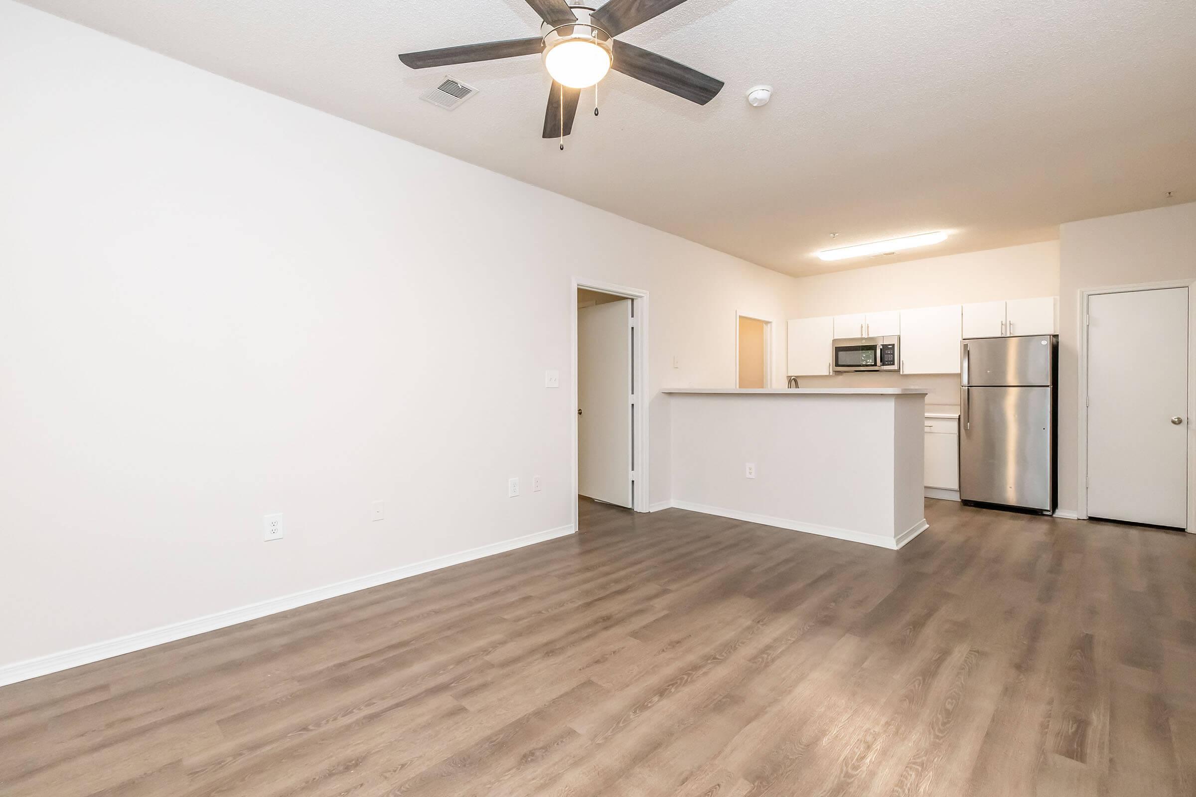 A spacious room featuring light-colored walls and wooden flooring, with a ceiling fan overhead. To the right, there is a kitchen area visible, equipped with modern appliances including a refrigerator and microwave. A doorway leads to another room, adding to the open layout.