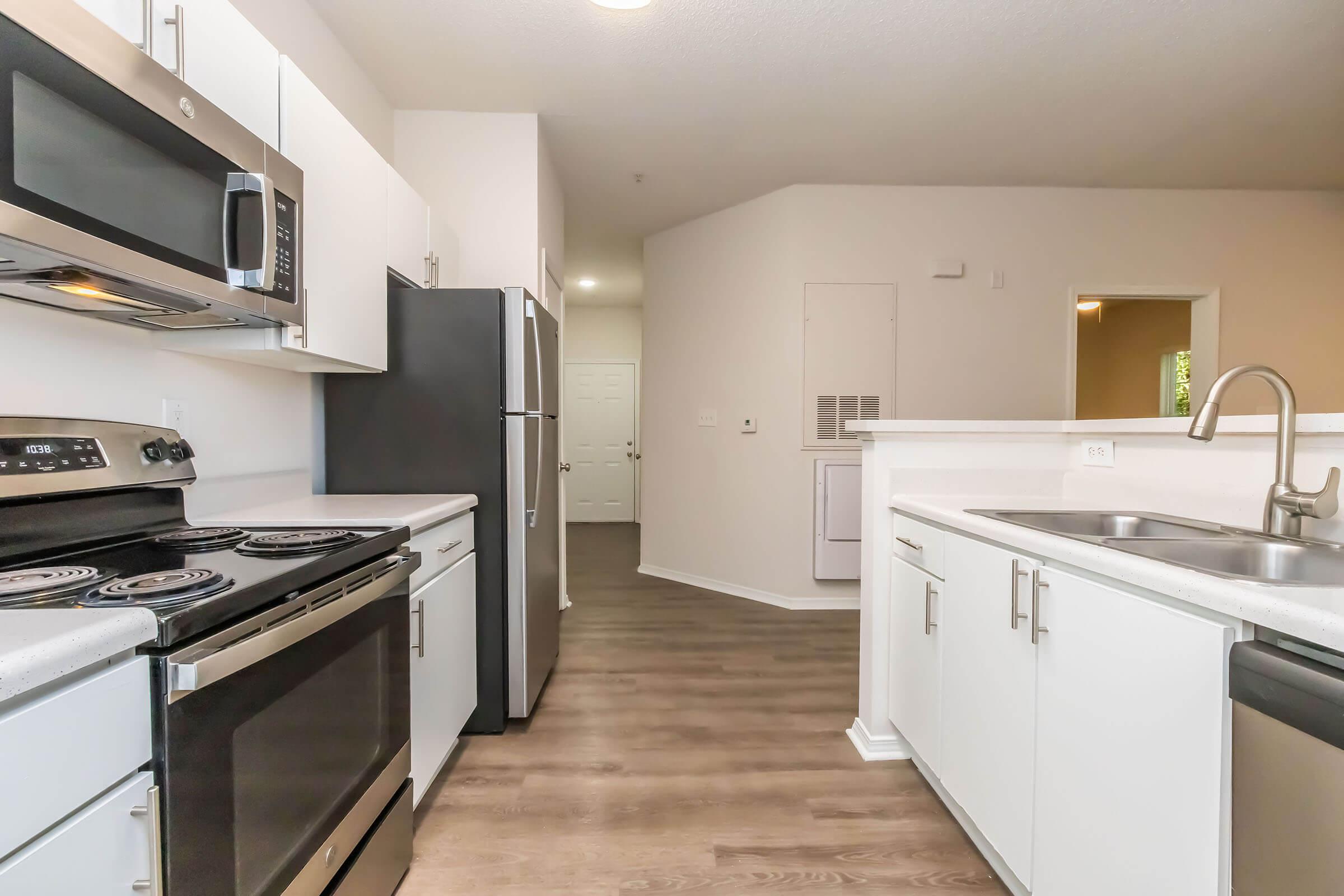 Modern kitchen featuring stainless steel appliances, including a microwave and oven, with white cabinets and a double sink. The kitchen has a neutral color scheme and wood-style flooring, with a view into an adjacent room. Natural light illuminates the space.