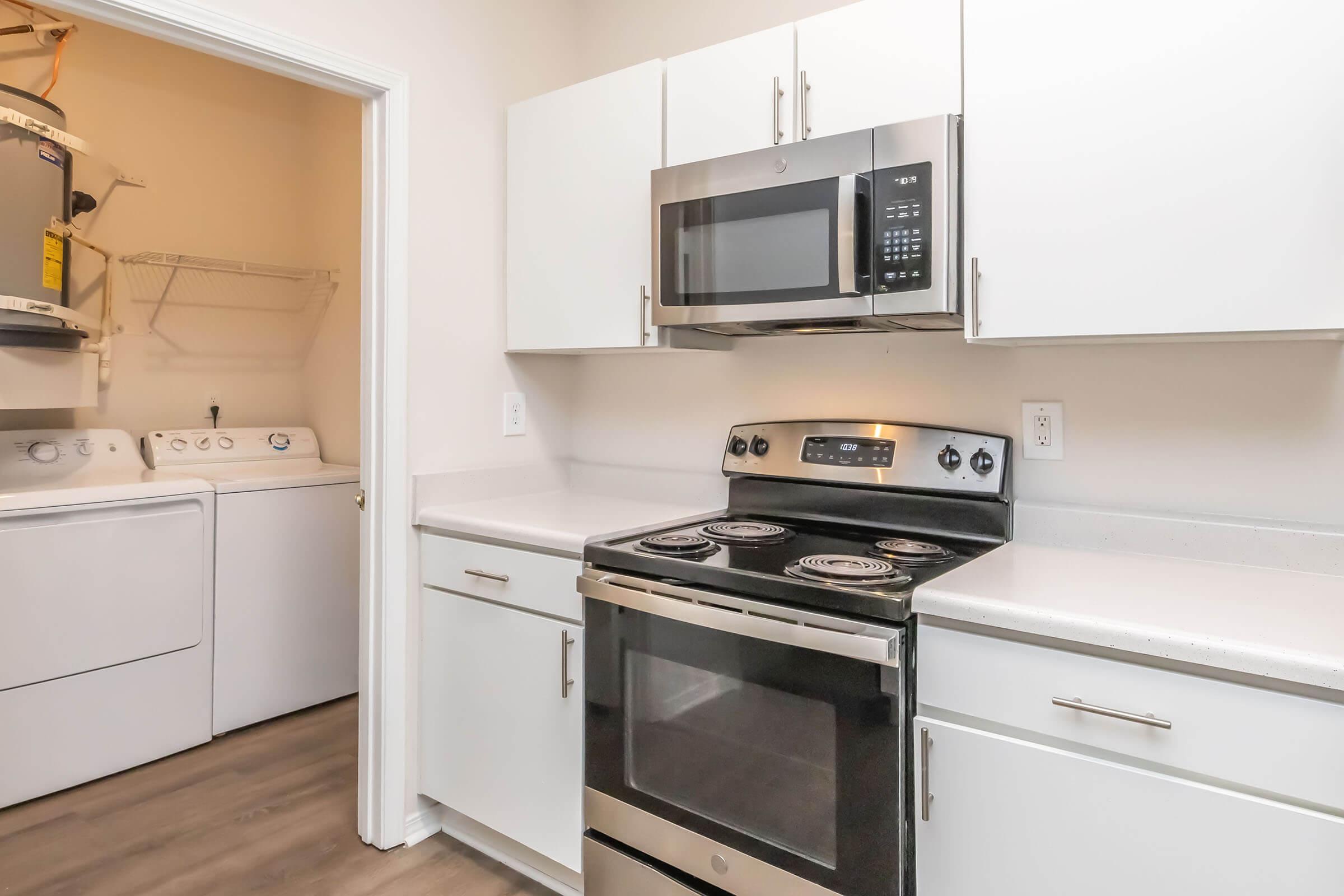A modern kitchen featuring a stainless steel oven and microwave, white cabinetry, and a clean countertop. To the left, a laundry area with a washing machine and dryer is visible, making efficient use of space in a compact design. The flooring is a light wood grain, enhancing the bright atmosphere.