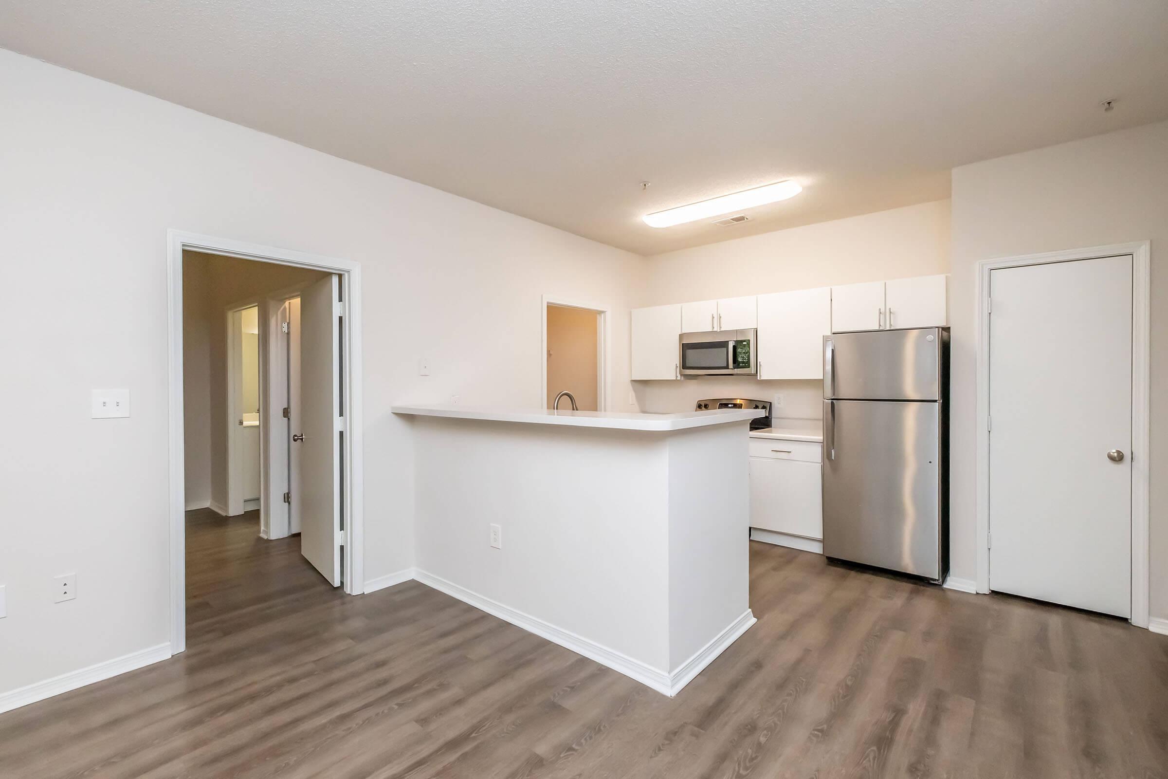 A modern, open-concept kitchen and living area with light wood flooring. The kitchen features a stainless steel refrigerator, stove, microwave, and white cabinetry. There are two doorways leading to other rooms, and the walls are painted in a neutral color, creating a bright and airy atmosphere.