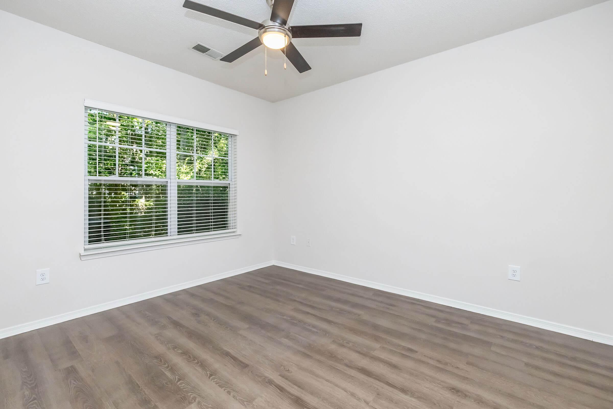 Empty room with light-colored walls and a fan on the ceiling. A window with white blinds allows natural light to enter, showcasing greenery outside. The floor is made of light wood, and the space is freshly painted and unfurnished, creating a minimalist atmosphere.