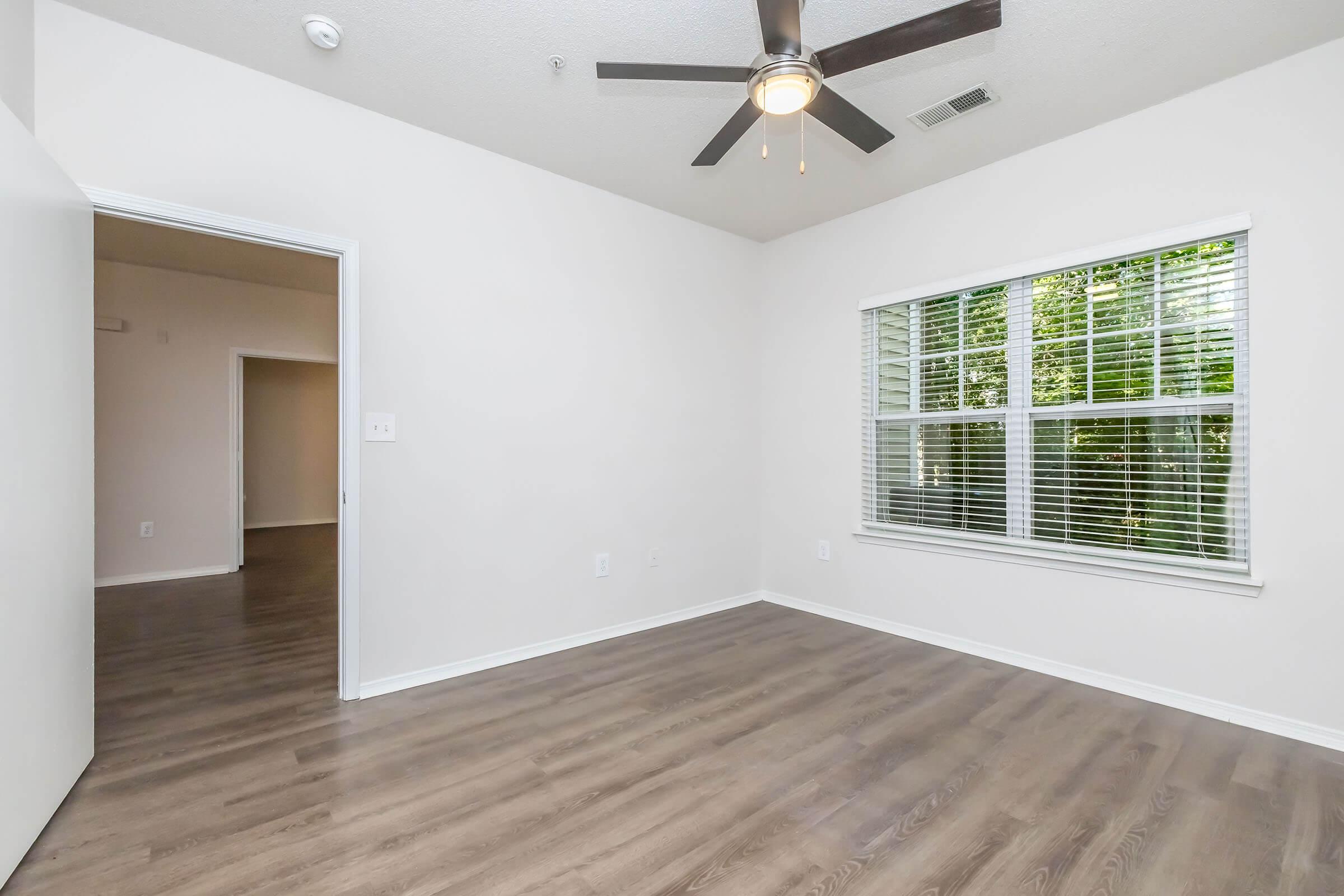 A bright, empty room with light-colored walls and a ceiling fan. The floor is a warm wood laminate. A window with blinds allows natural light to enter, revealing trees outside. There's an open doorway leading to another room, creating an inviting and spacious feel.