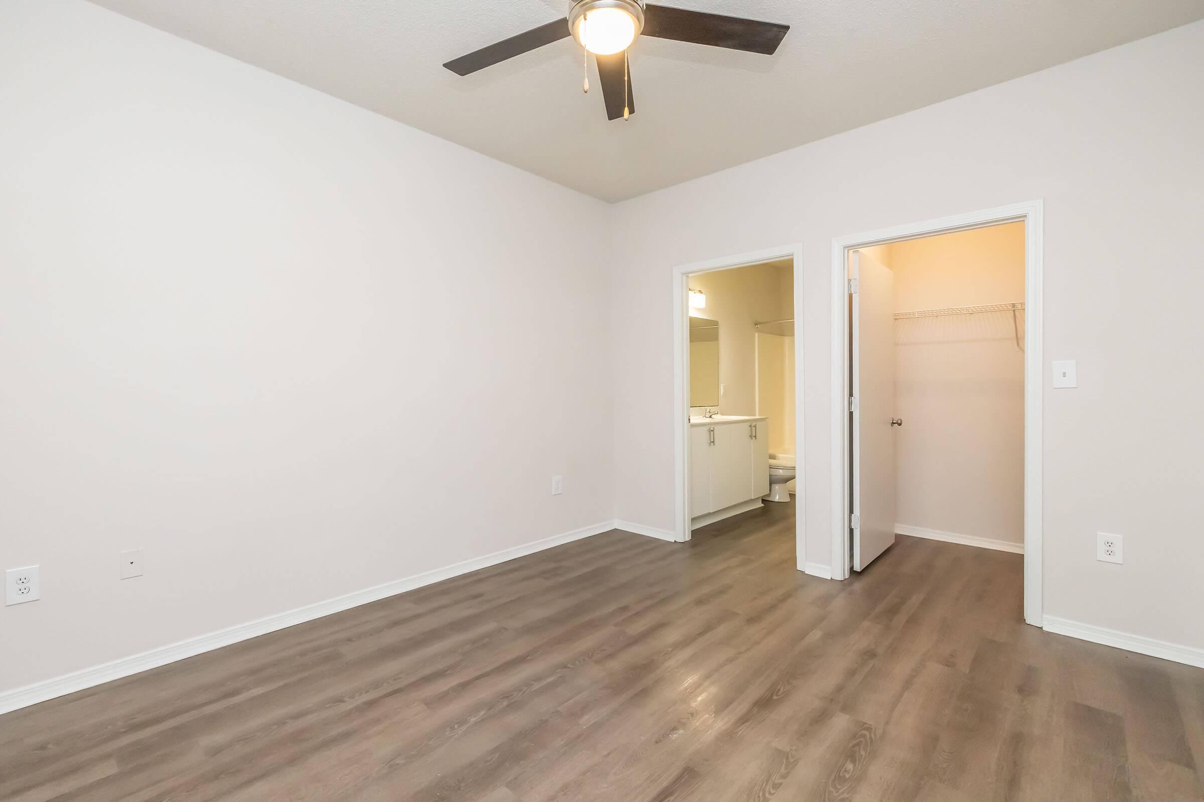 A view of a spacious, empty room with light-colored walls and hardwood flooring. A ceiling fan is visible above. Two doorways lead to other areas, one likely a bathroom and the other a closet. The room is bright and uncluttered, creating an open, airy atmosphere.