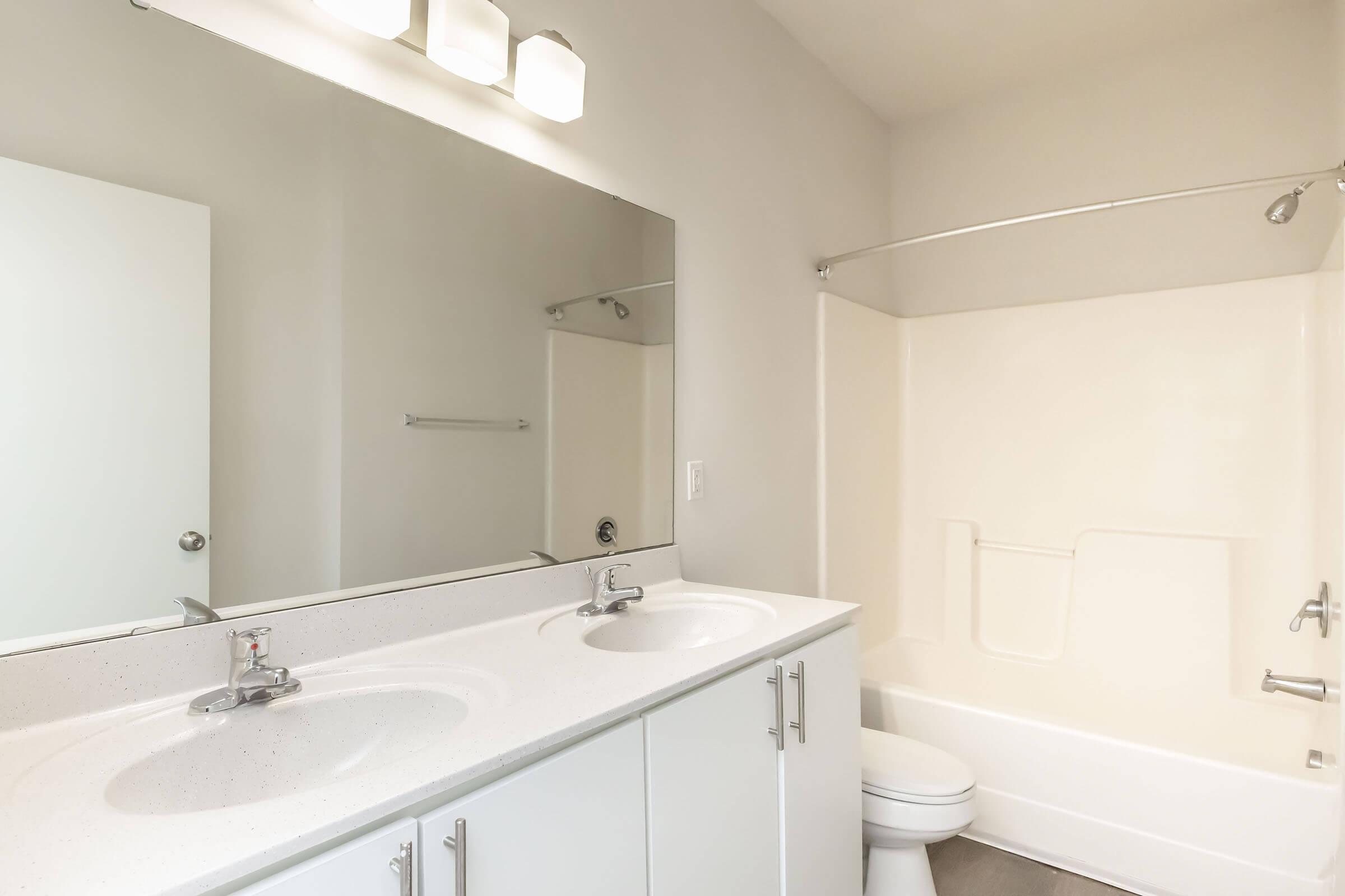 A clean, modern bathroom featuring a double sink vanity with two faucets, a large mirror, and bright lighting. The room includes a separate toilet area and a bath/shower combination with a smooth, white finish. The walls are light-colored, creating a bright and spacious atmosphere.