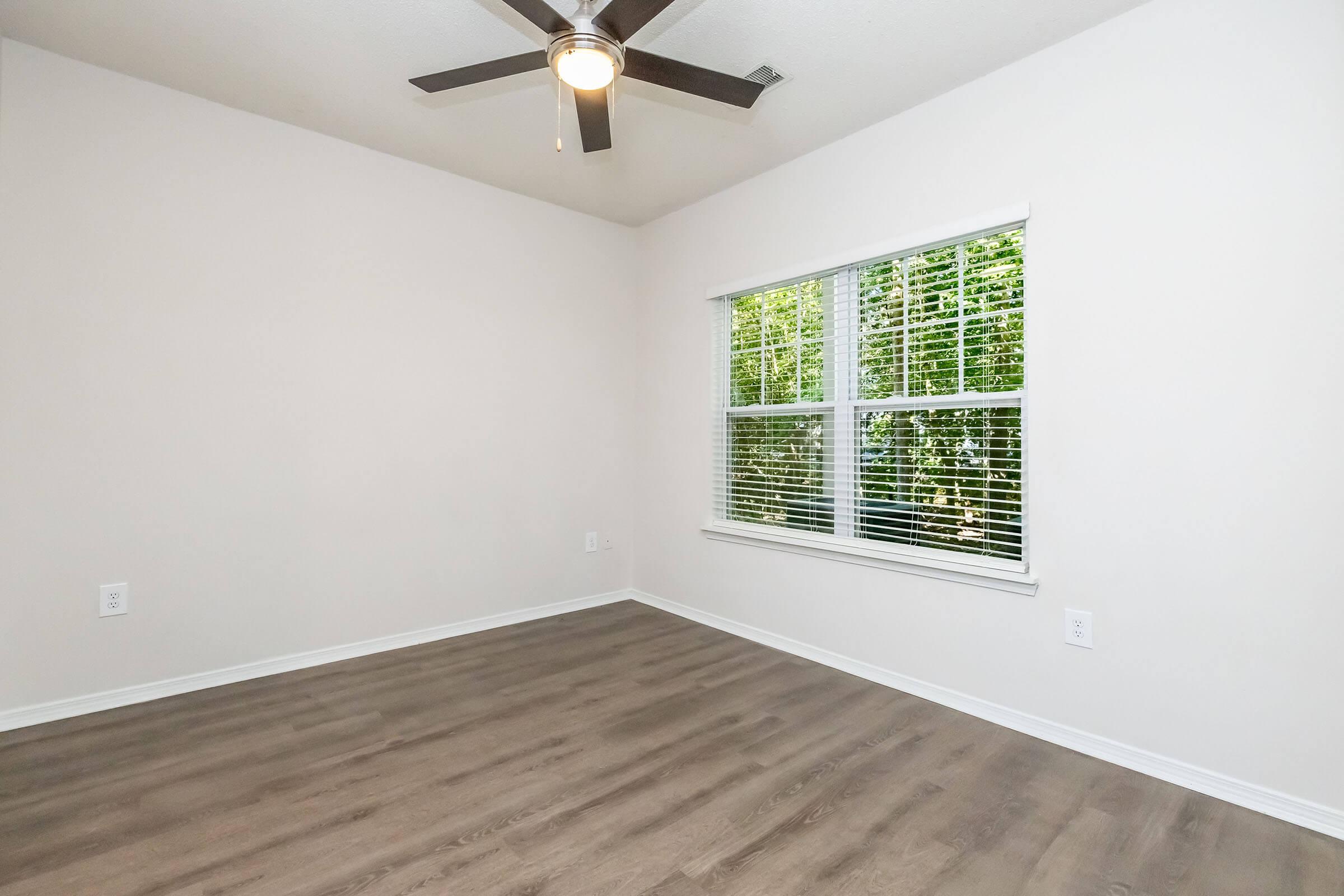 A bright and empty room with light-colored walls and a ceiling fan. It features a large window with blinds that overlooks greenery outside. The floor is covered with light wooden laminate, creating a clean and minimalist appearance.