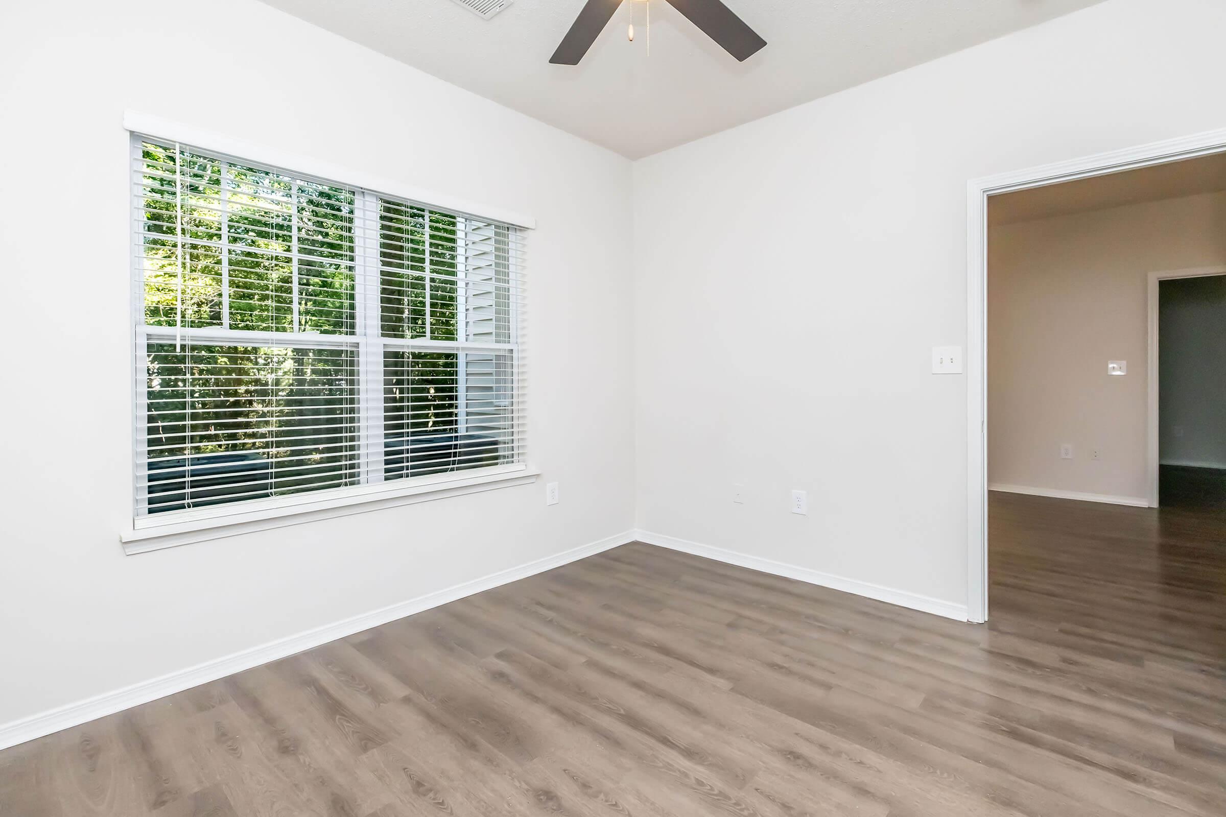 A bright, empty room with light-colored walls and a ceiling fan. A large window with blinds allows natural light to enter, showcasing a view of greenery outside. The floor is made of wood or laminate, giving the space a warm feel. Another room is visible in the background through an open doorway.