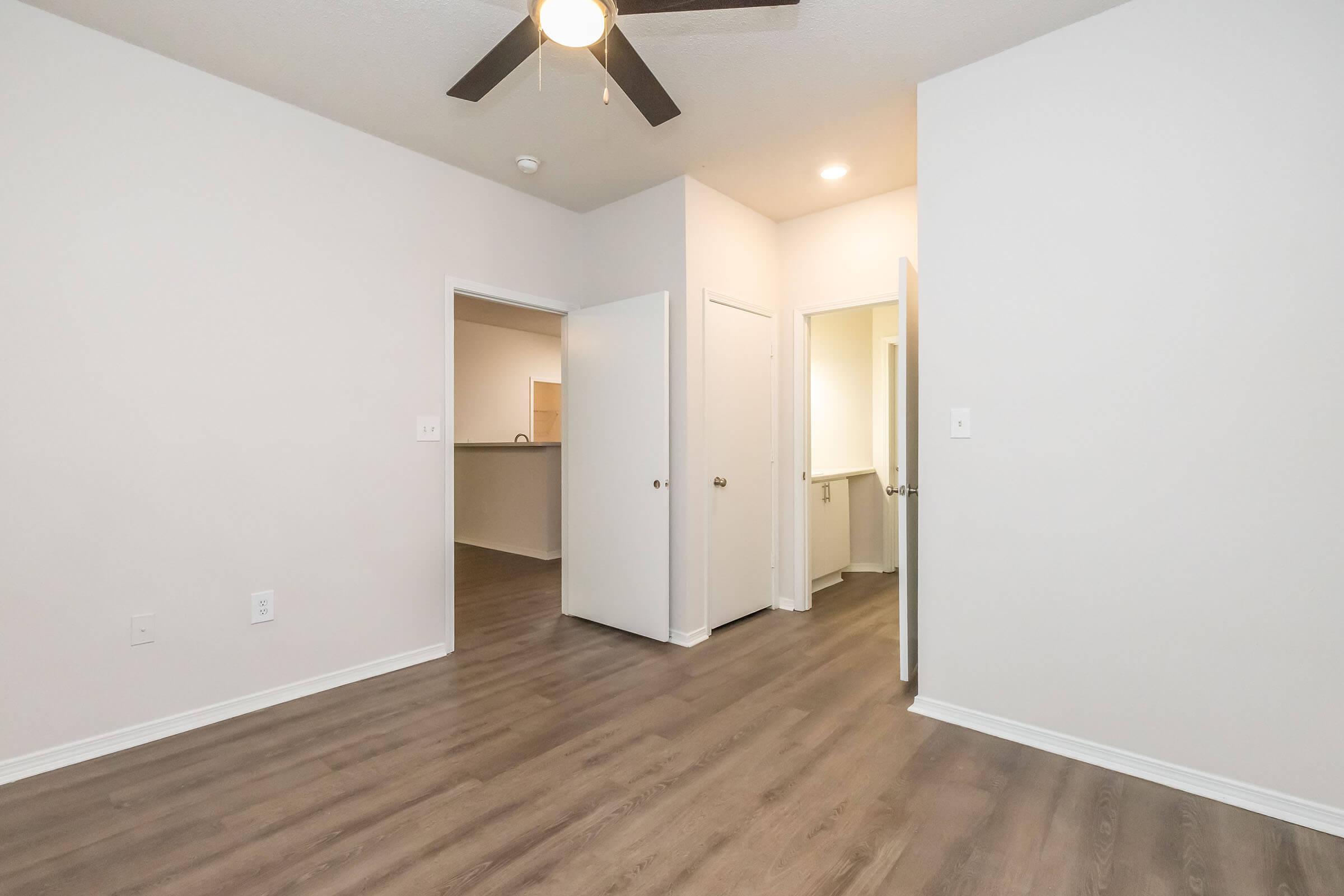 A small, empty room with light-colored walls and brown laminate flooring. A ceiling fan is visible above. There are doorways leading to other spaces, including a visible kitchen area in the background. The room has natural light and a clean, modern appearance.