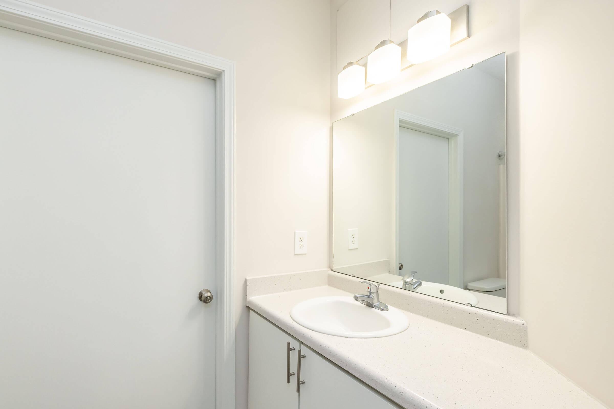 A bright restroom featuring a simple white sink with a faucet, a large mirror above the sink, and three modern light fixtures. The walls are painted in a light color, and a closed door is visible on the left side. The countertop is a smooth white surface, creating a clean and minimalistic aesthetic.
