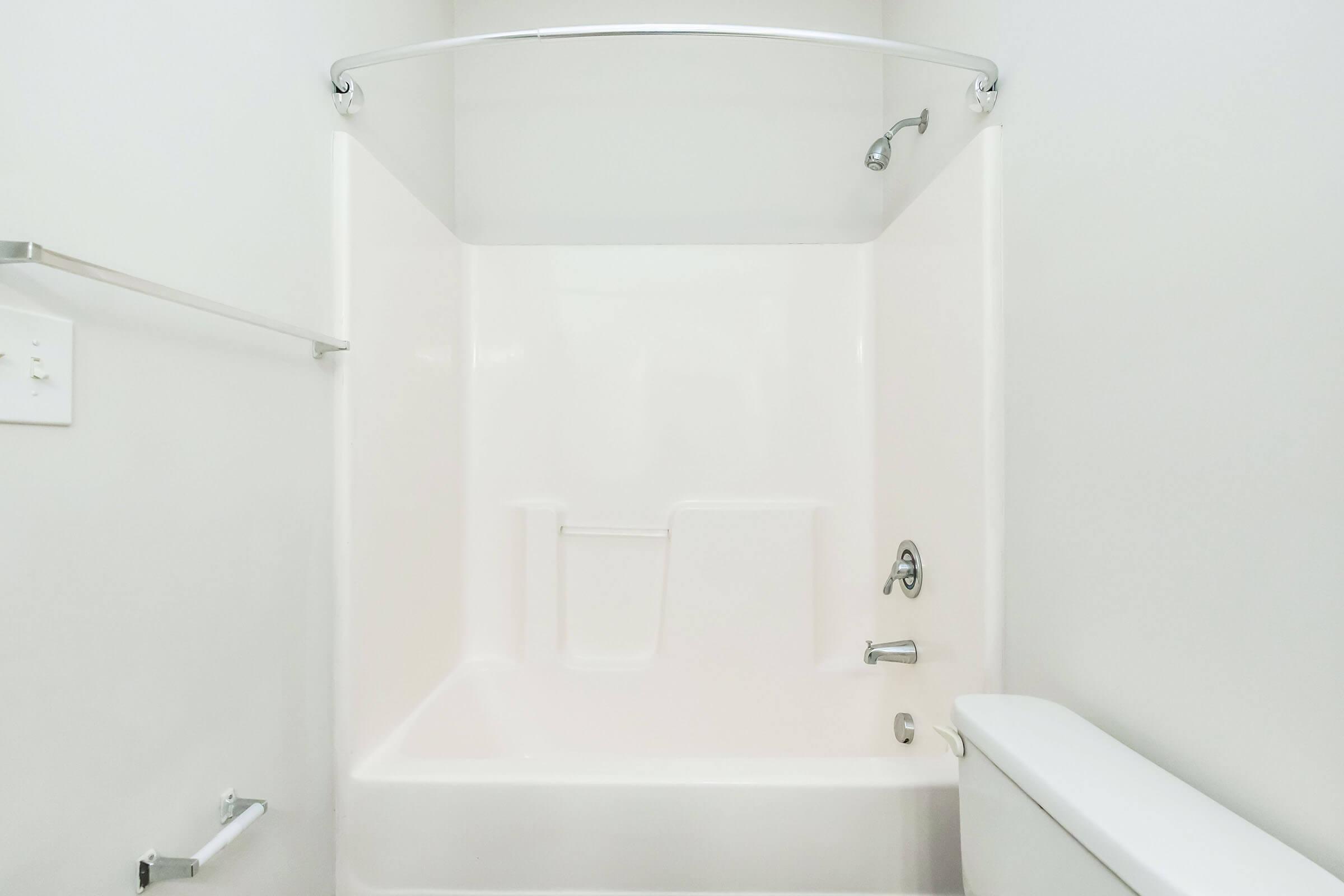 A clean, minimalistic bathroom featuring a white acrylic bathtub with a curved shower rod, a single showerhead, and a toilet nearby. The walls are plain and light-colored, contributing to a bright and spacious feel.
