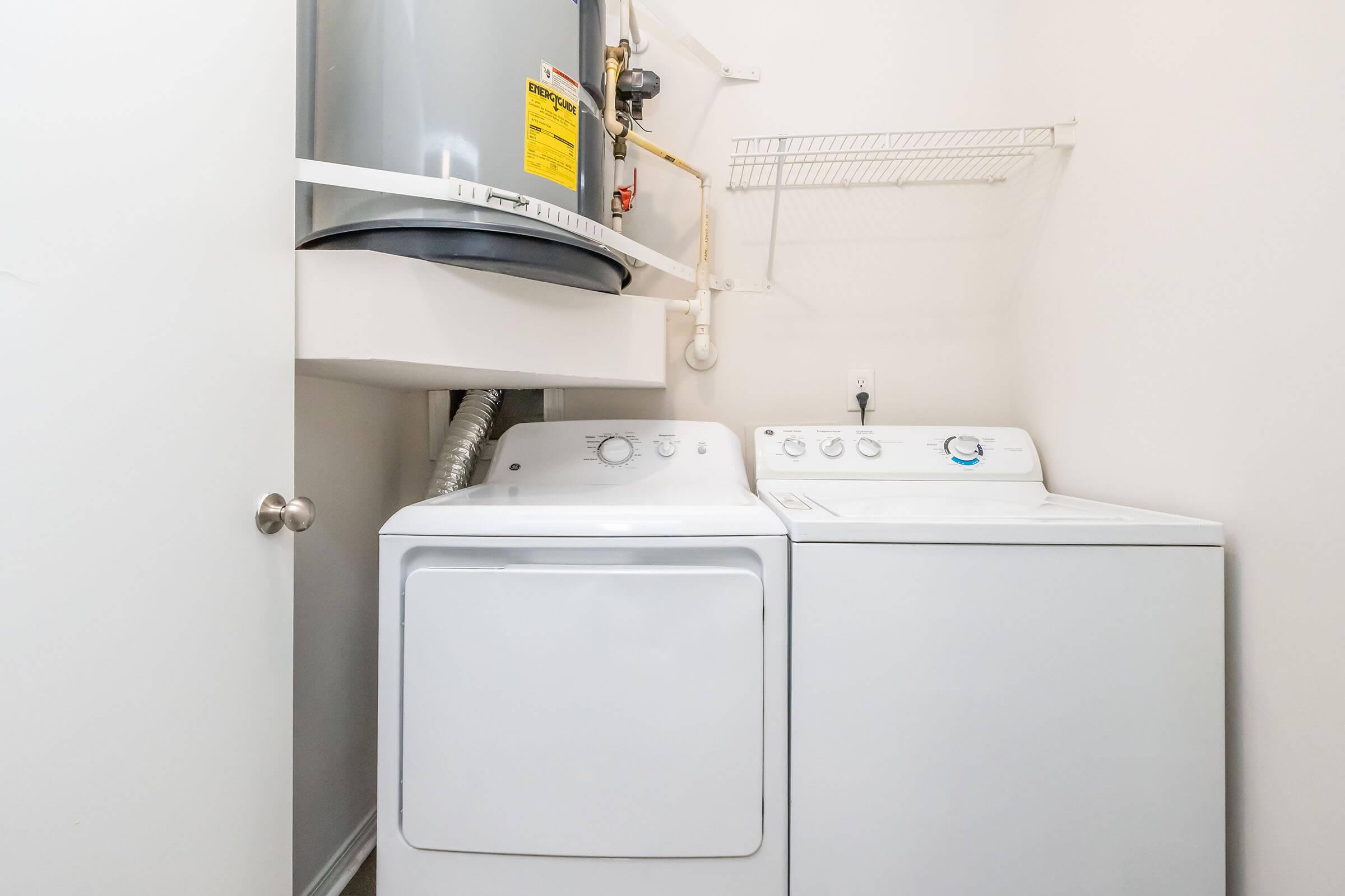 A laundry room featuring a washing machine and a dryer side by side. Above them, there's a water heater mounted on the wall. A shelf hangs above for storage, and the walls are painted a light color, creating a clean and functional space.