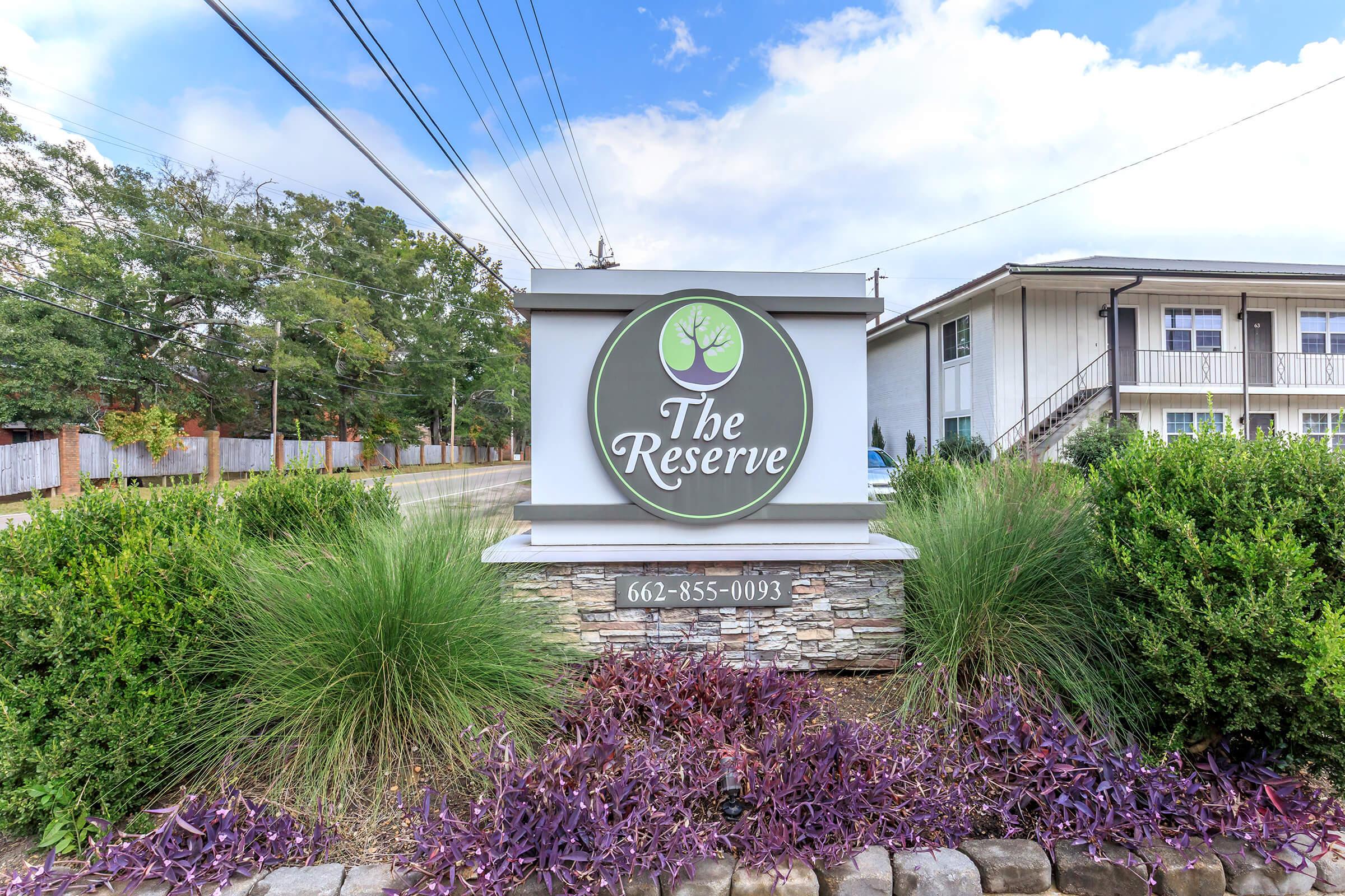 Sign for "The Reserve" with a circular logo featuring a tree. The sign is surrounded by landscaping that includes green grass and purple plants. In the background, there are power lines and a white building with multiple units. The contact number is displayed as 662-855-0093.