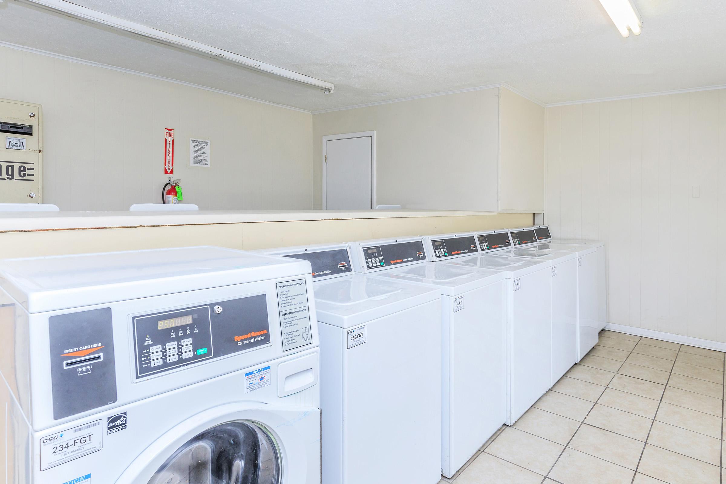 A clean laundry room featuring several white washing machines lined up against a wall. The floor is tiled, and overhead fluorescent lighting illuminates the space. There are instructions and safety signs posted nearby, and a door can be seen at the far end of the room.