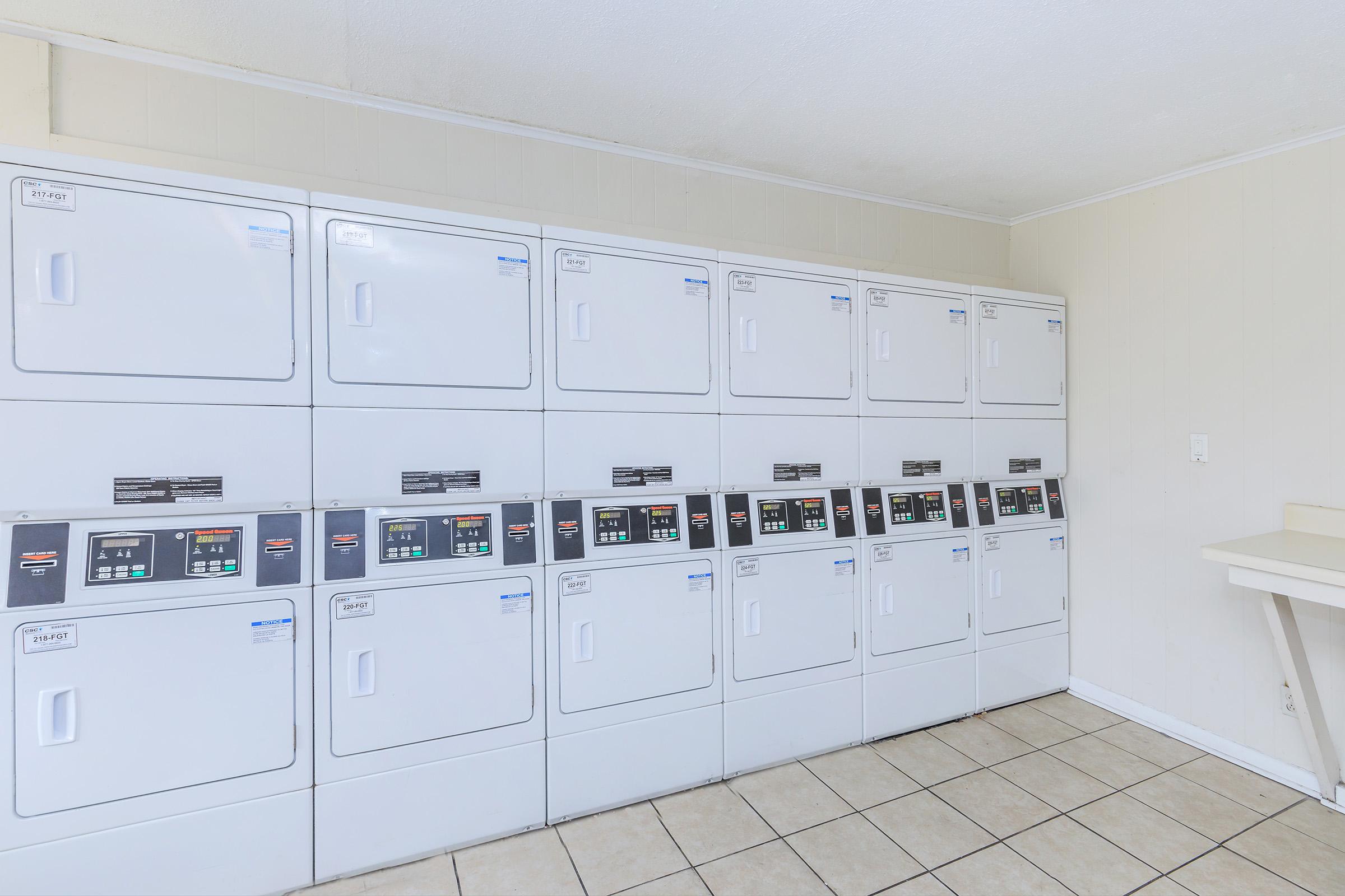 A clean, well-lit laundry room featuring a row of stacked white washing machines and dryers. The machines have digital control panels and are lined up against a light-colored wall. A small counter is visible on the right side, and the floor is tiled.