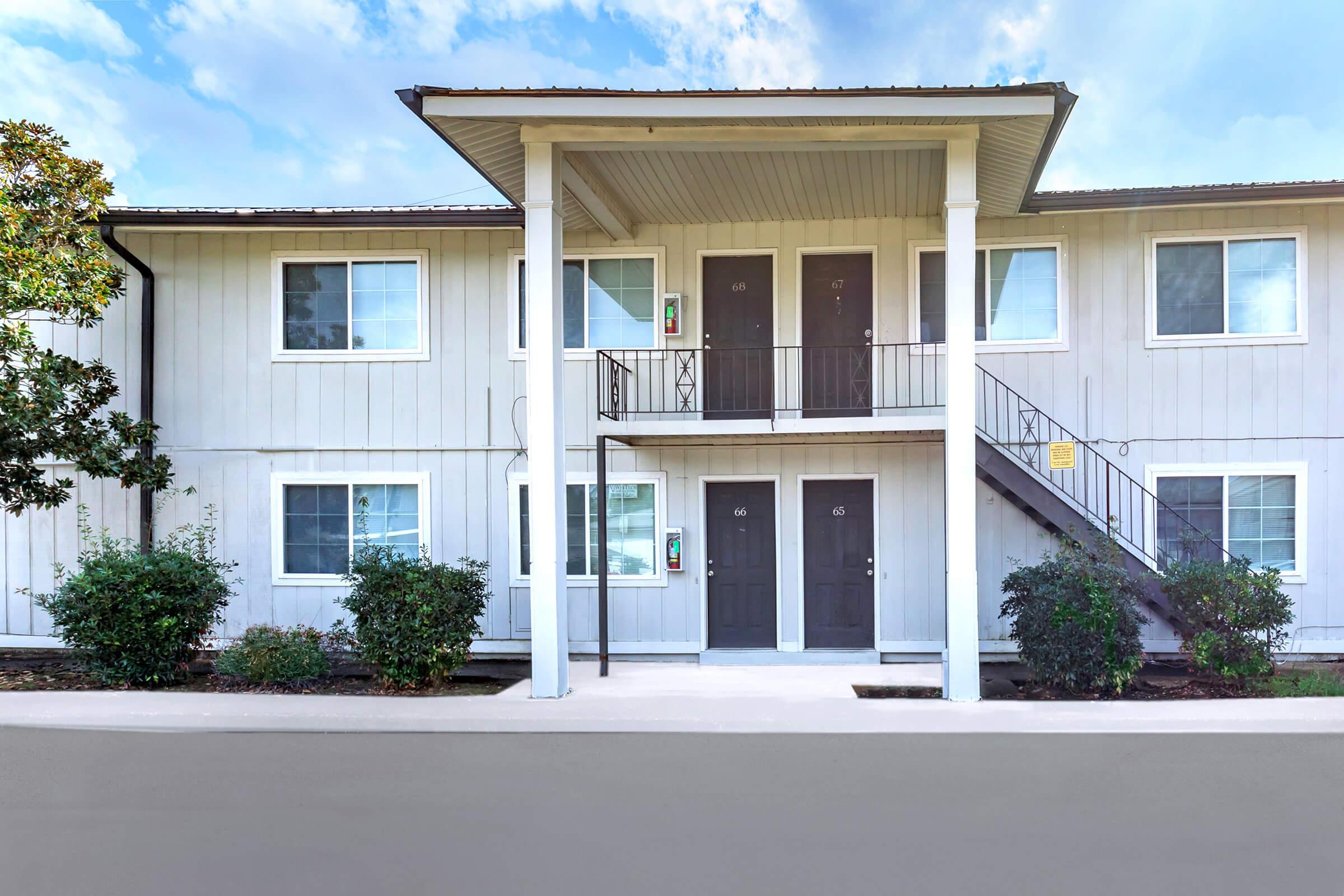 Two-story residential building with a light gray exterior. It features a central staircase leading to two sets of doors on each floor. Lush green bushes are planted in front, and the area is well-maintained, creating a welcoming atmosphere. The sky above is clear with soft clouds.