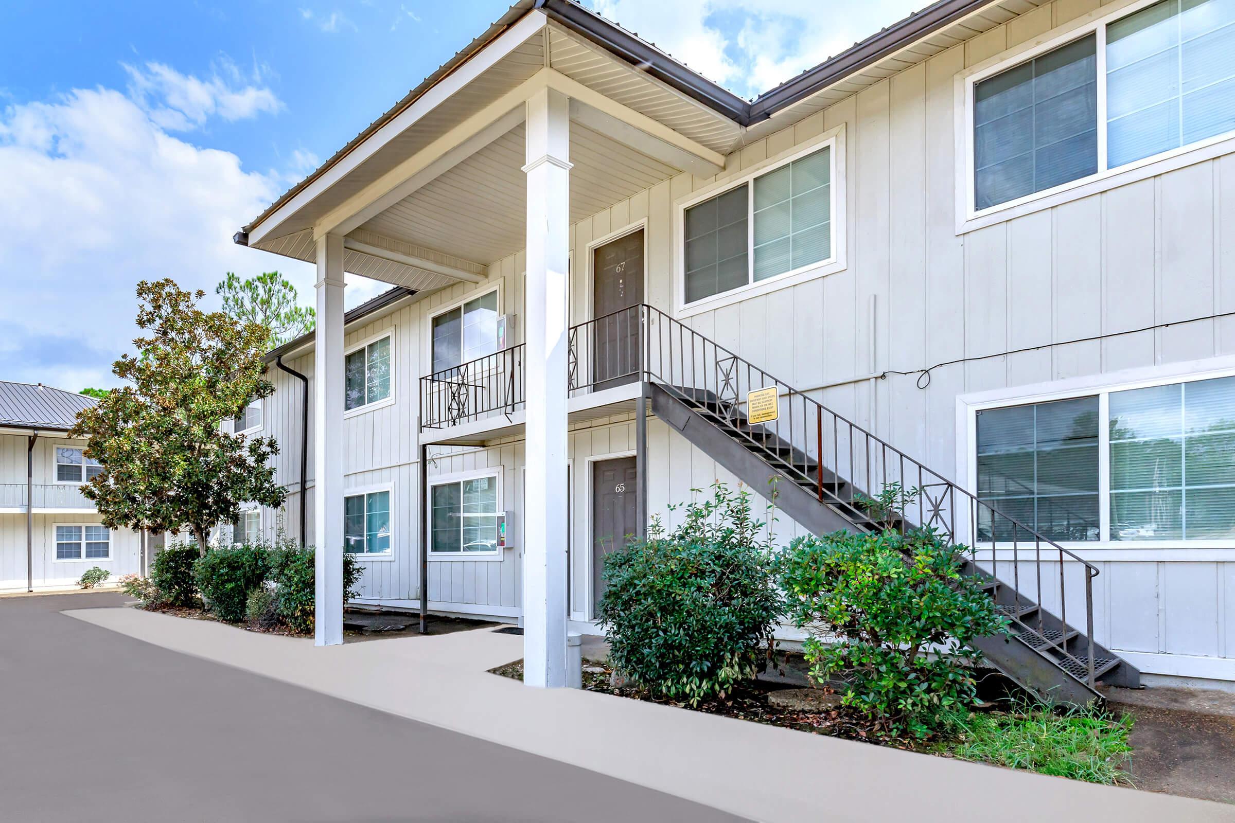 A two-story apartment building with a white exterior. Stairs lead to the second floor, where there are multiple doors. Surrounding the building are neatly trimmed shrubs and trees. The sky is partly cloudy, and the scene conveys a quiet residential atmosphere.