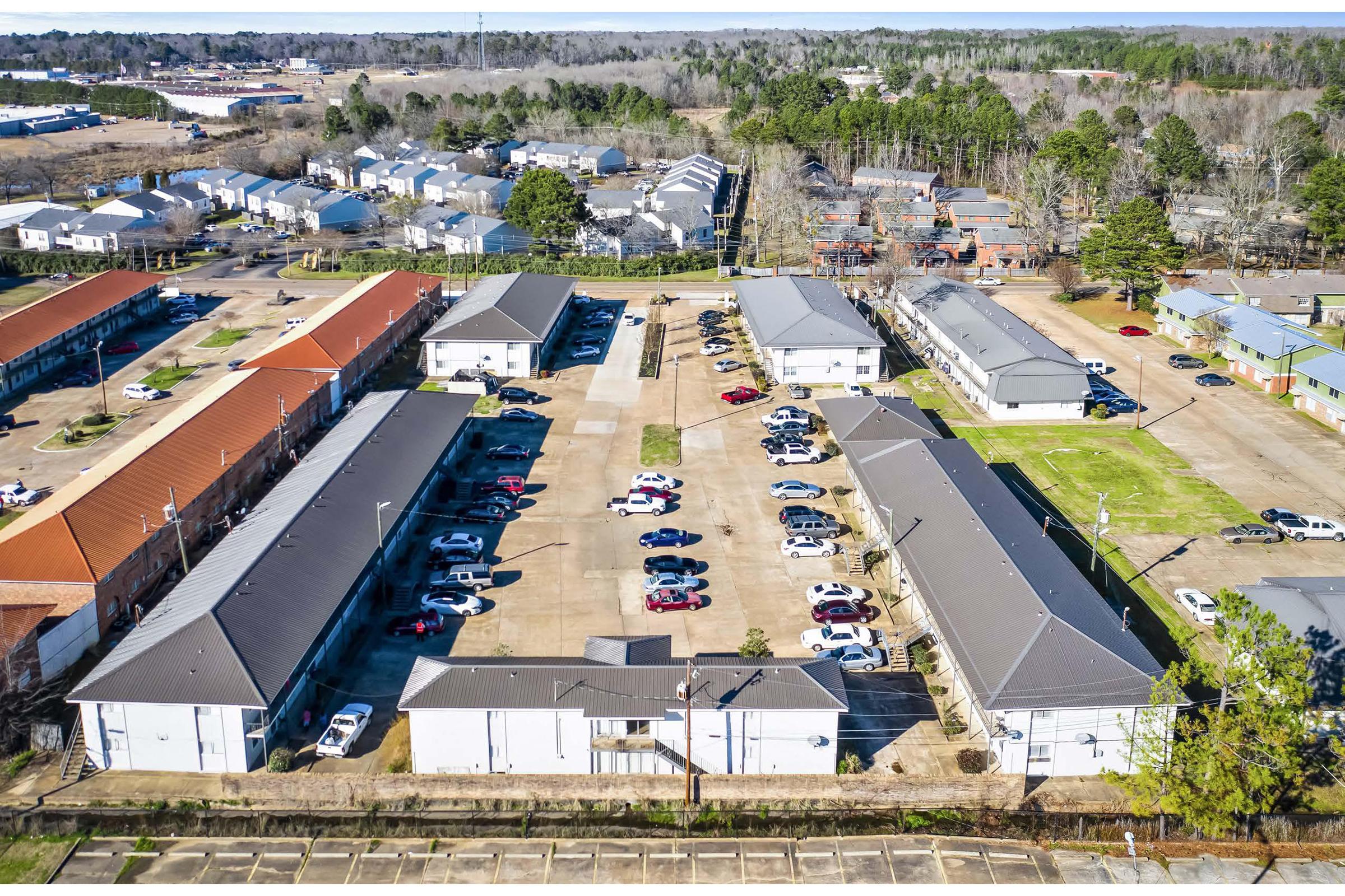 Aerial view of a residential complex featuring multiple buildings with gray roofs, surrounded by parking spaces for vehicles. The layout shows several rows of apartments, with some cars parked in designated areas. Nearby, there are trees and additional residential structures in the background.