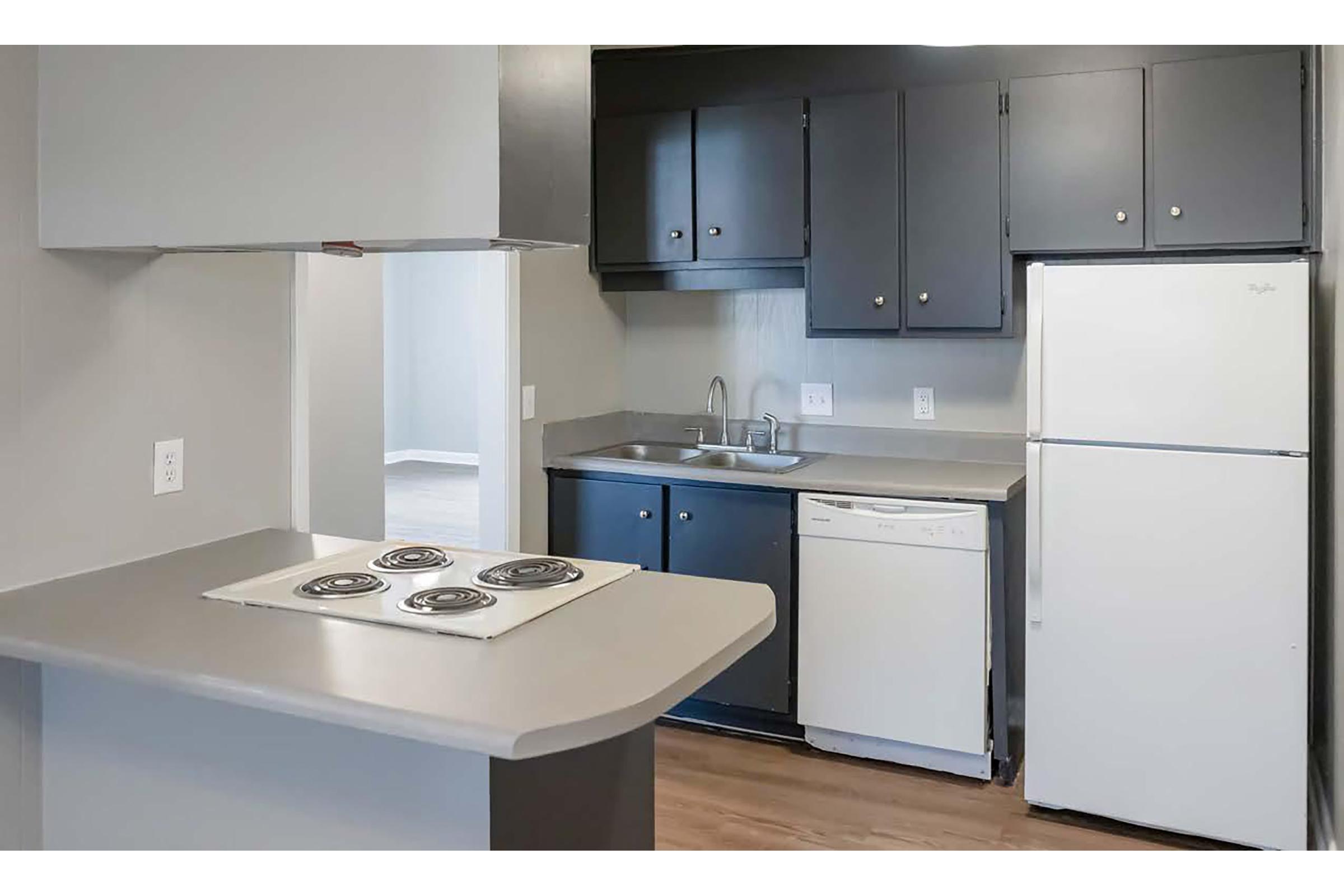 A modern kitchen featuring dark gray cabinets, a light-colored countertop, a white refrigerator, a built-in dishwasher, and an electric stove with four burners. The space is well-lit with neutral colors and wood flooring, creating a functional and inviting atmosphere.