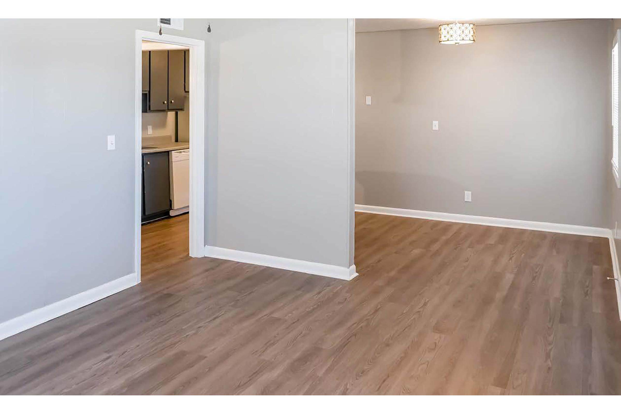 Interior view of a modern, empty room with light gray walls and wooden laminate flooring. A doorway leads to a kitchen area on the left, and a window is on the right side, allowing natural light to enter. A small light fixture is visible on the ceiling, enhancing the bright ambiance of the space.