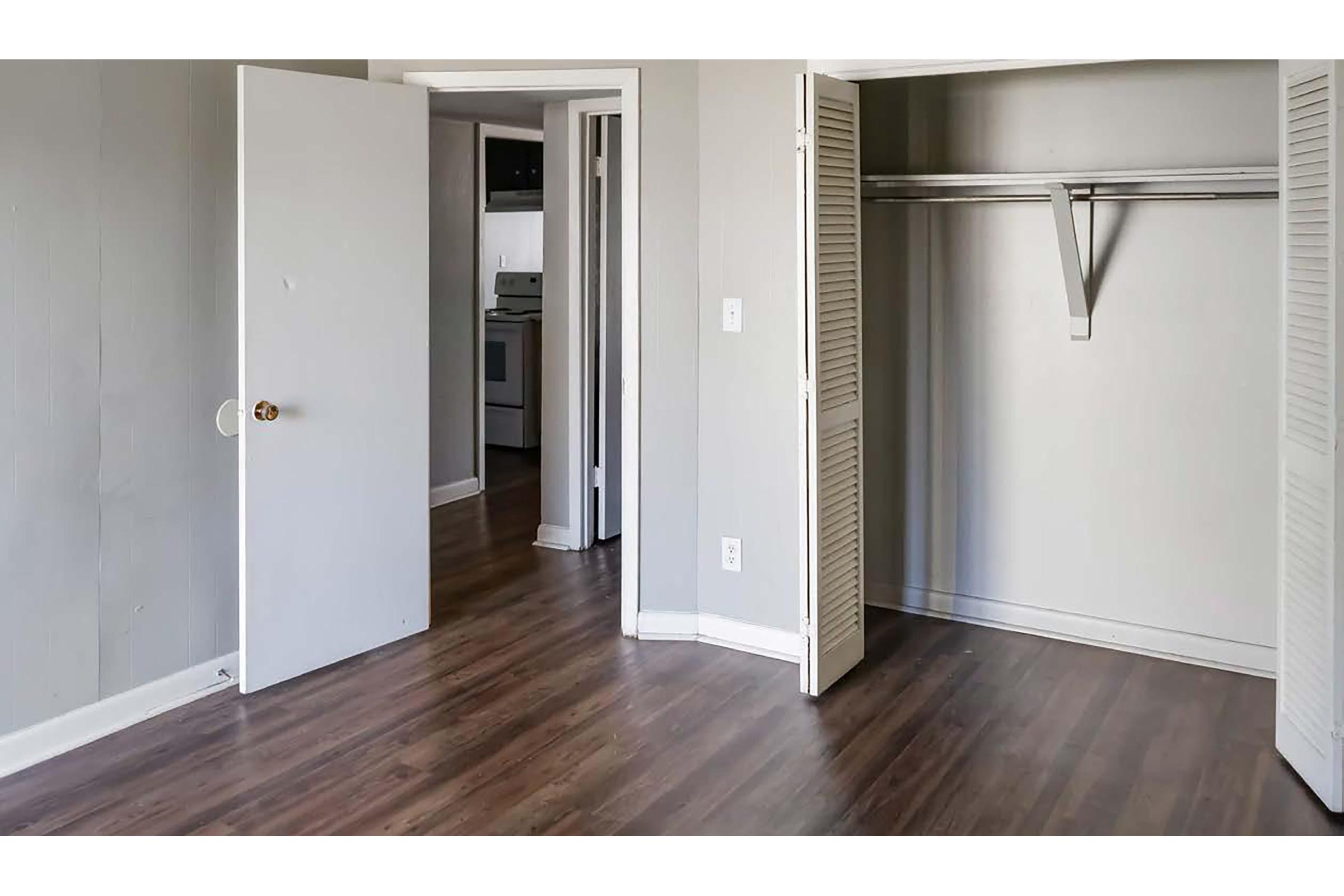 A partially furnished room with light gray walls and brown wooden flooring. The image shows an open closet with a hanging rack and a door leading to another room, likely a kitchen. The space appears bright and inviting, suitable for various uses.