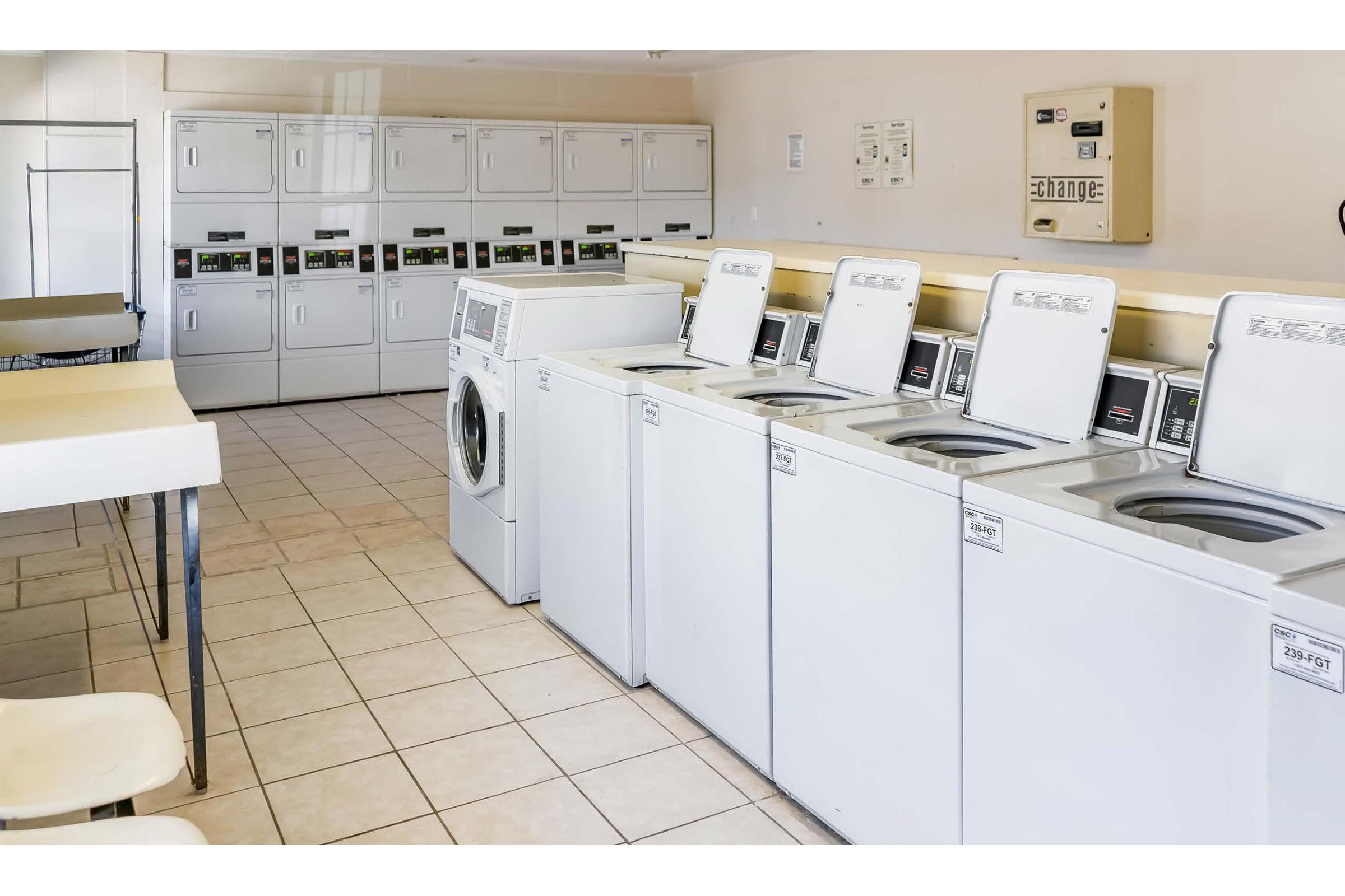 A clean, bright laundry room featuring several white washing machines and dryers arranged in a row, with a folding table nearby. The walls are light-colored, and there are multiple laundry machines against the back wall. The space is organized and well-maintained, suitable for communal laundry use.