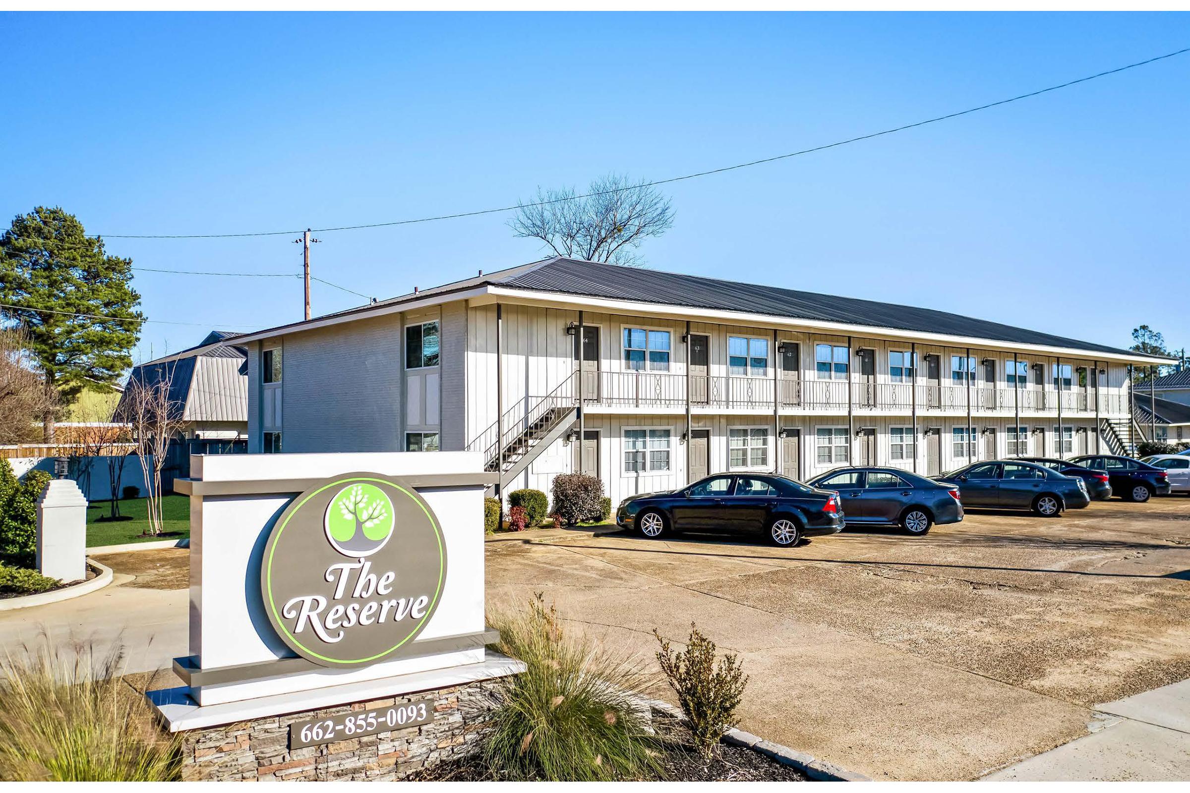 A modern multi-story apartment building with exterior staircases and balconies, surrounded by parked cars. A sign in front reads "The Reserve" with a tree logo, indicating the property's name. The landscape features greenery and a clear blue sky in the background.