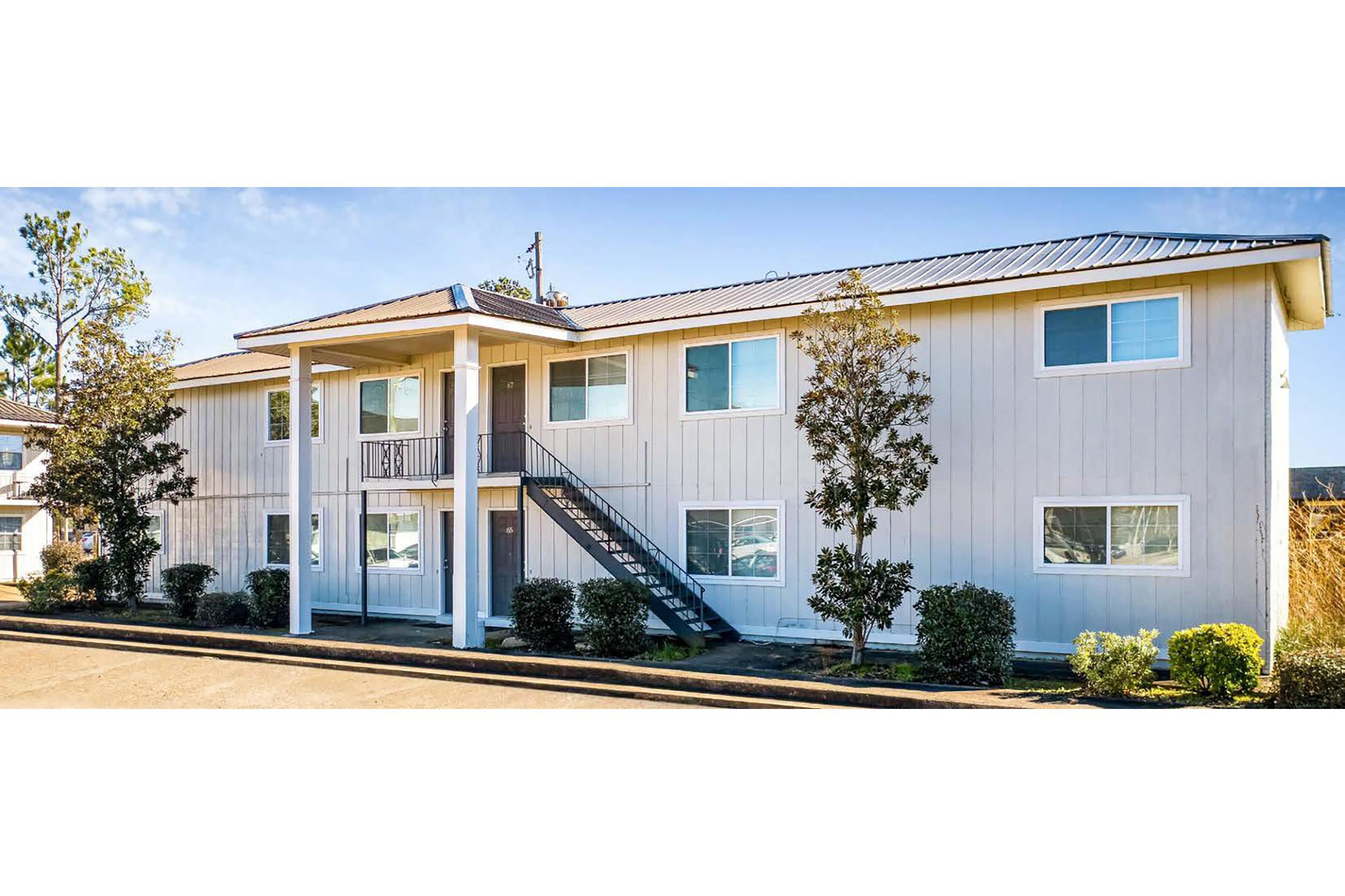 A two-story apartment building with a light gray exterior, white trim, and a metal roof. The building features a central staircase and multiple windows. Surrounding the building are small trees and shrubs, with a clear blue sky in the background. The pavement leads up to the entrance area.