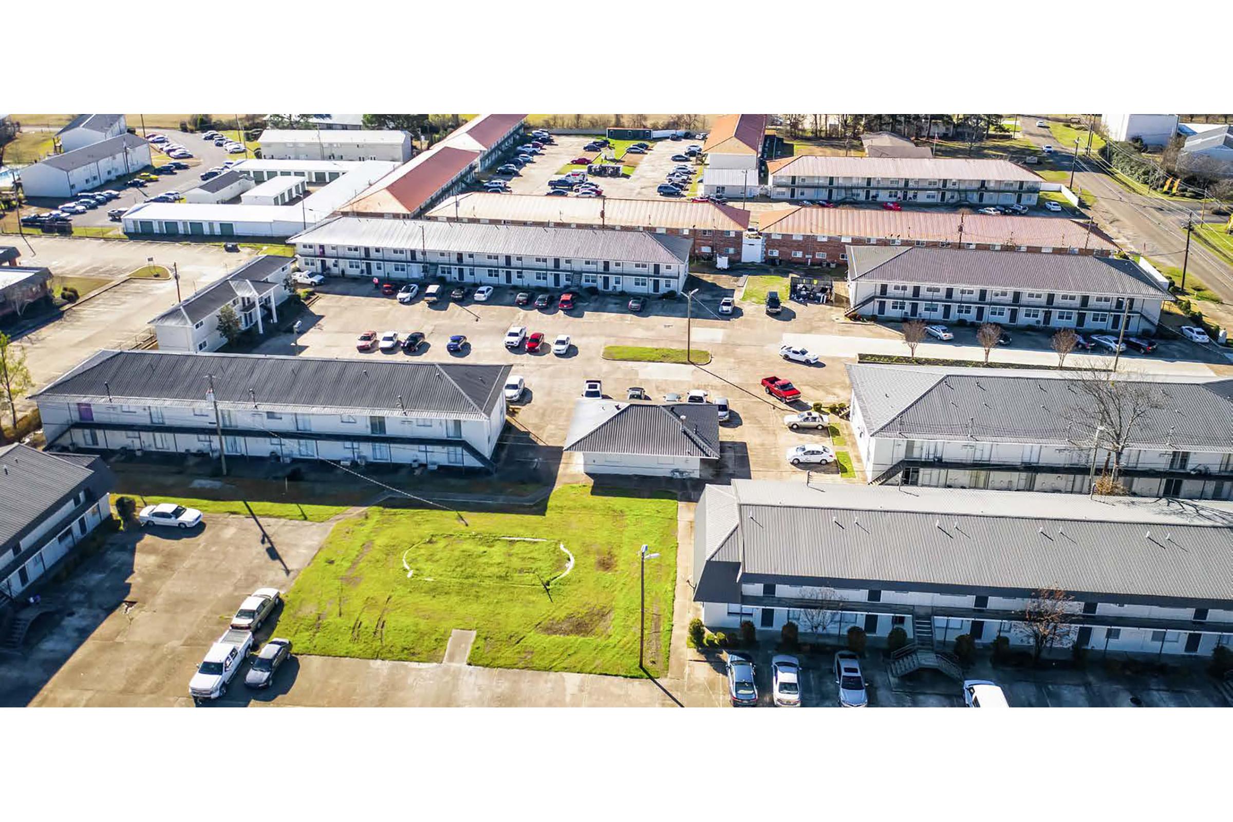 An aerial view of a parking area surrounded by low-rise buildings. Several vehicles are parked in designated spaces, with green grass visible in the center. The layout includes multiple units connected by walkways, with additional parking visible in the background.