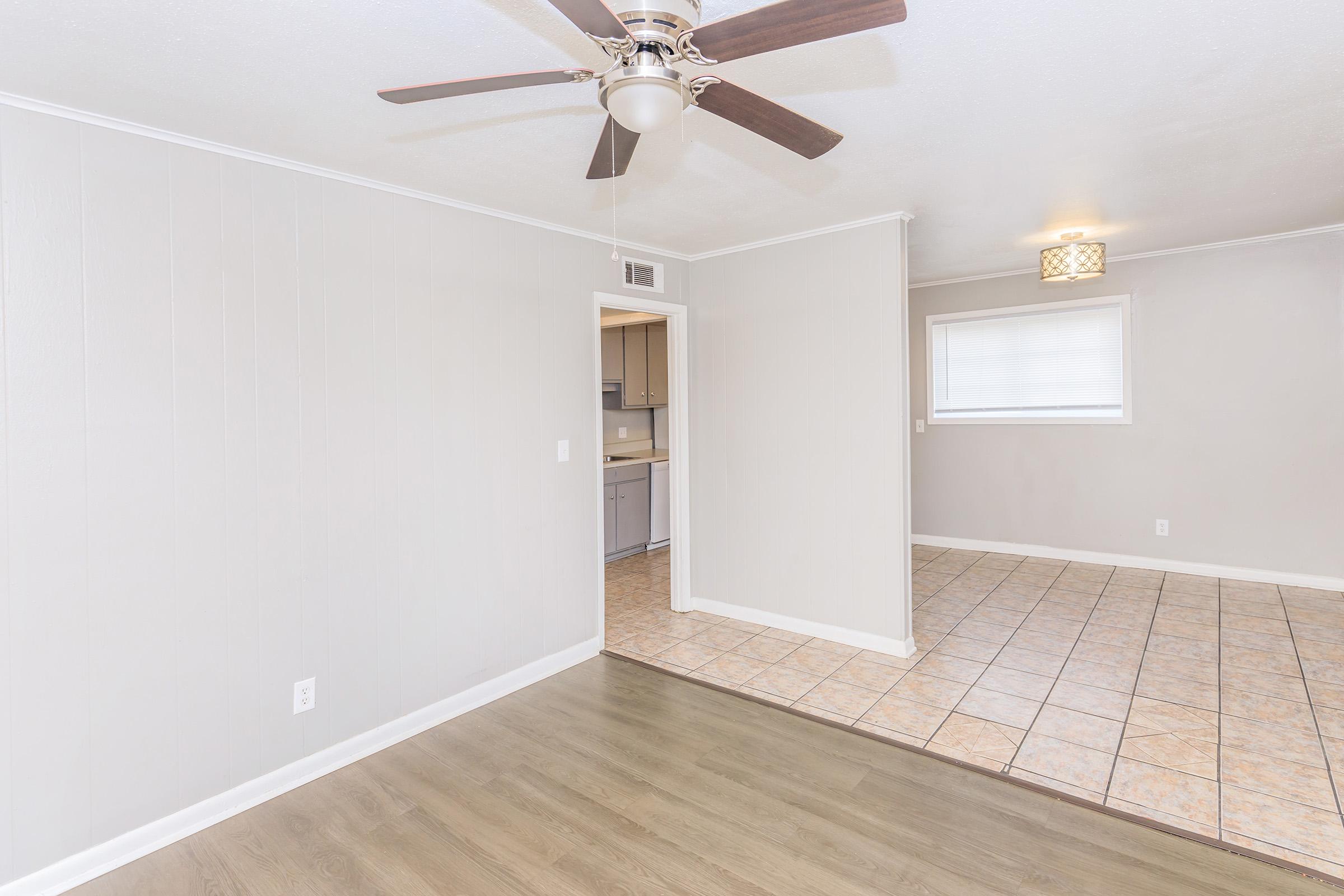 A spacious, well-lit living area with a ceiling fan and a mix of tile and wood flooring. The room features light gray walls and an open layout leading to a kitchen area in the background, with natural light coming from a window.