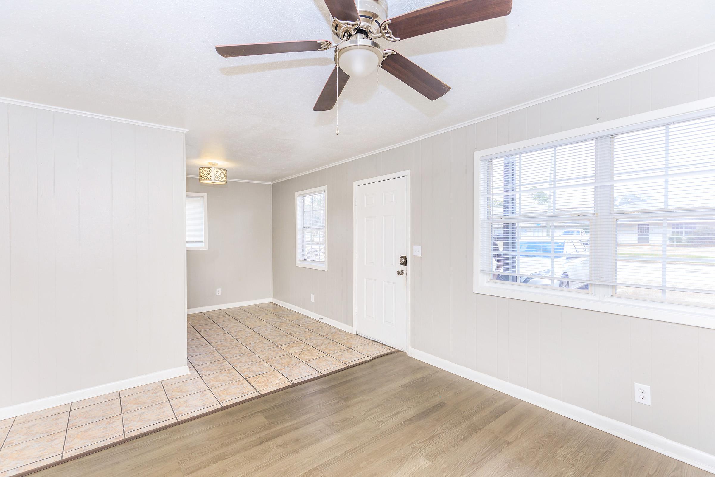 Interior view of a home featuring a beige wall, a light-colored tile floor, and dark wood flooring. A ceiling fan is mounted on the ceiling, and there are large windows allowing natural light. The entryway is visible, with a door leading outside.