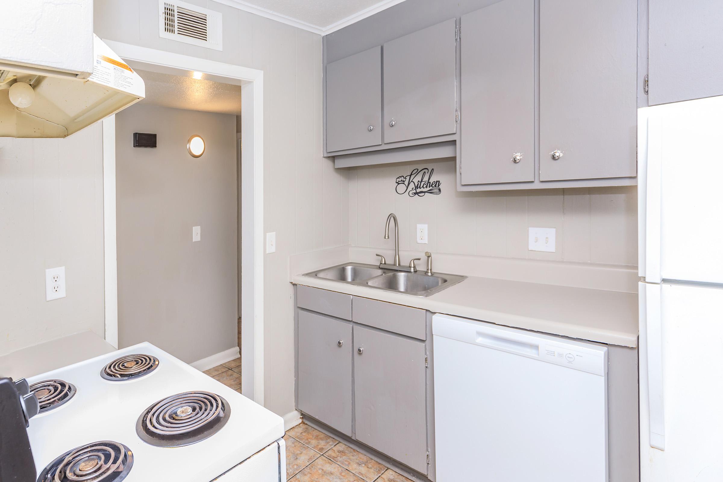 Compact kitchen featuring gray cabinetry, a double sink, white appliances including a refrigerator and dishwasher, and a stove with four burners. The walls are light gray, and there's a tile floor. A sign with "Kitchen" is displayed on the wall.