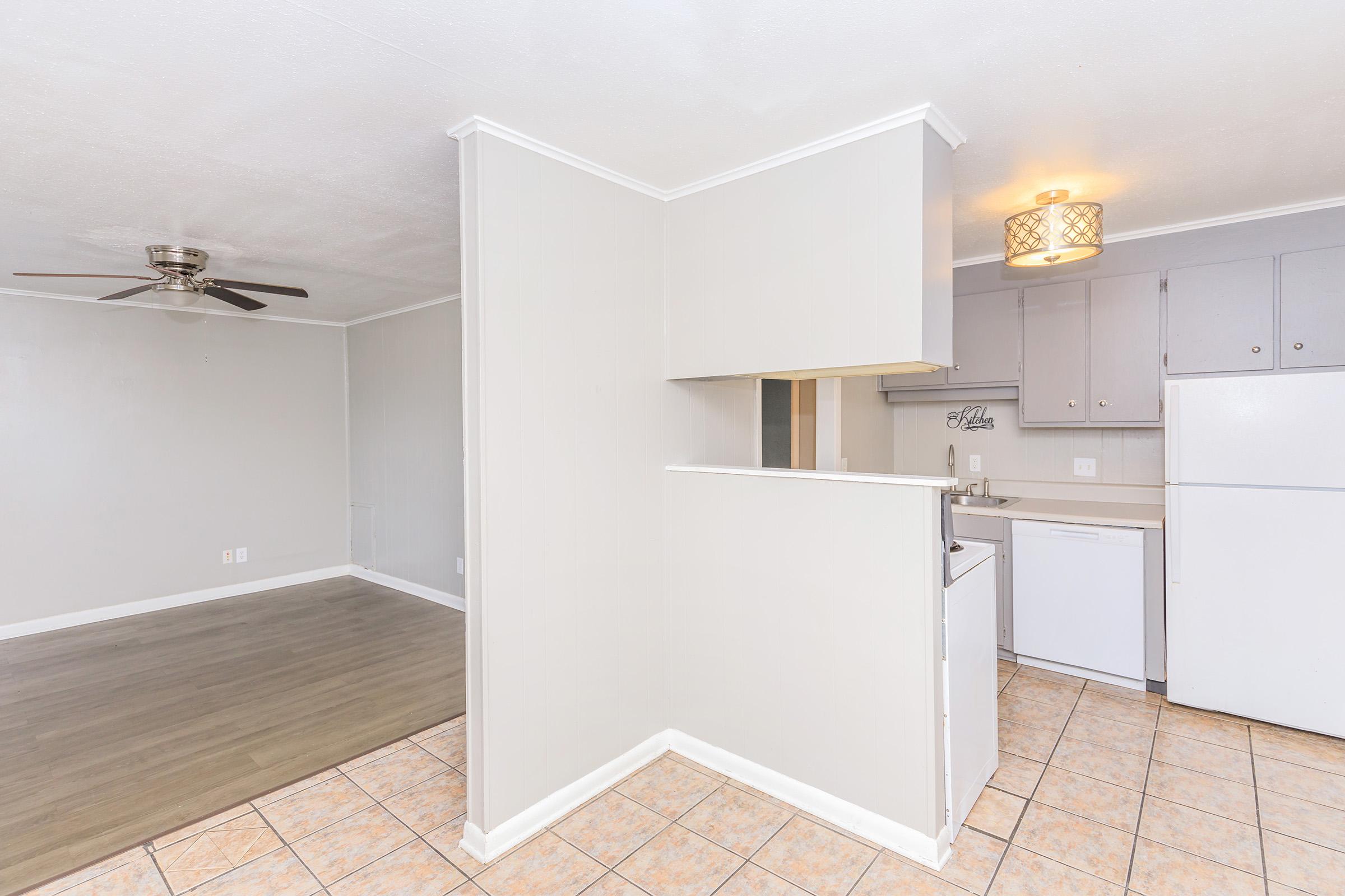 A view of an open living space featuring a kitchen area with light gray cabinets, a refrigerator, and a dishwasher. The adjacent room is empty with a light-colored floor, and there's a ceiling fan visible. The walls are painted in neutral tones, contributing to a bright and spacious atmosphere.