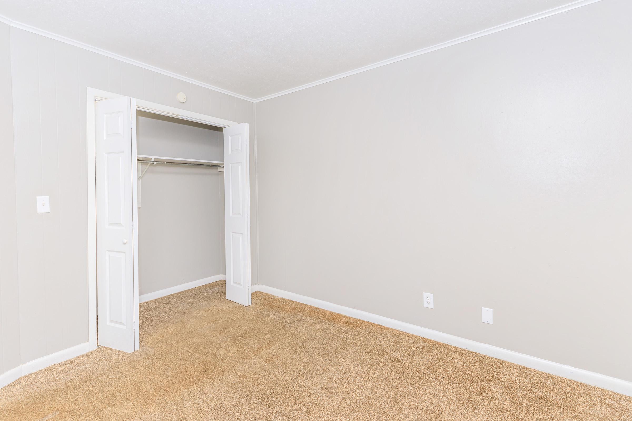 A clean, empty room with light gray walls and plush beige carpeting. There is a closet with double doors on the left side, which is open and contains a hanging rail. The room has no furniture, giving it a spacious and minimalist appearance.