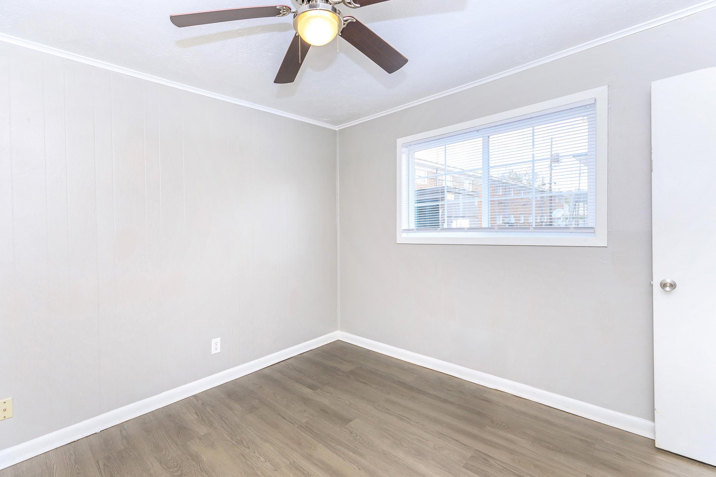 A small room with light gray walls and a window. The floor is wood-like, and there's a ceiling fan with a light fixture. A white door is visible on the right side, and the room appears to be unoccupied and well-lit.