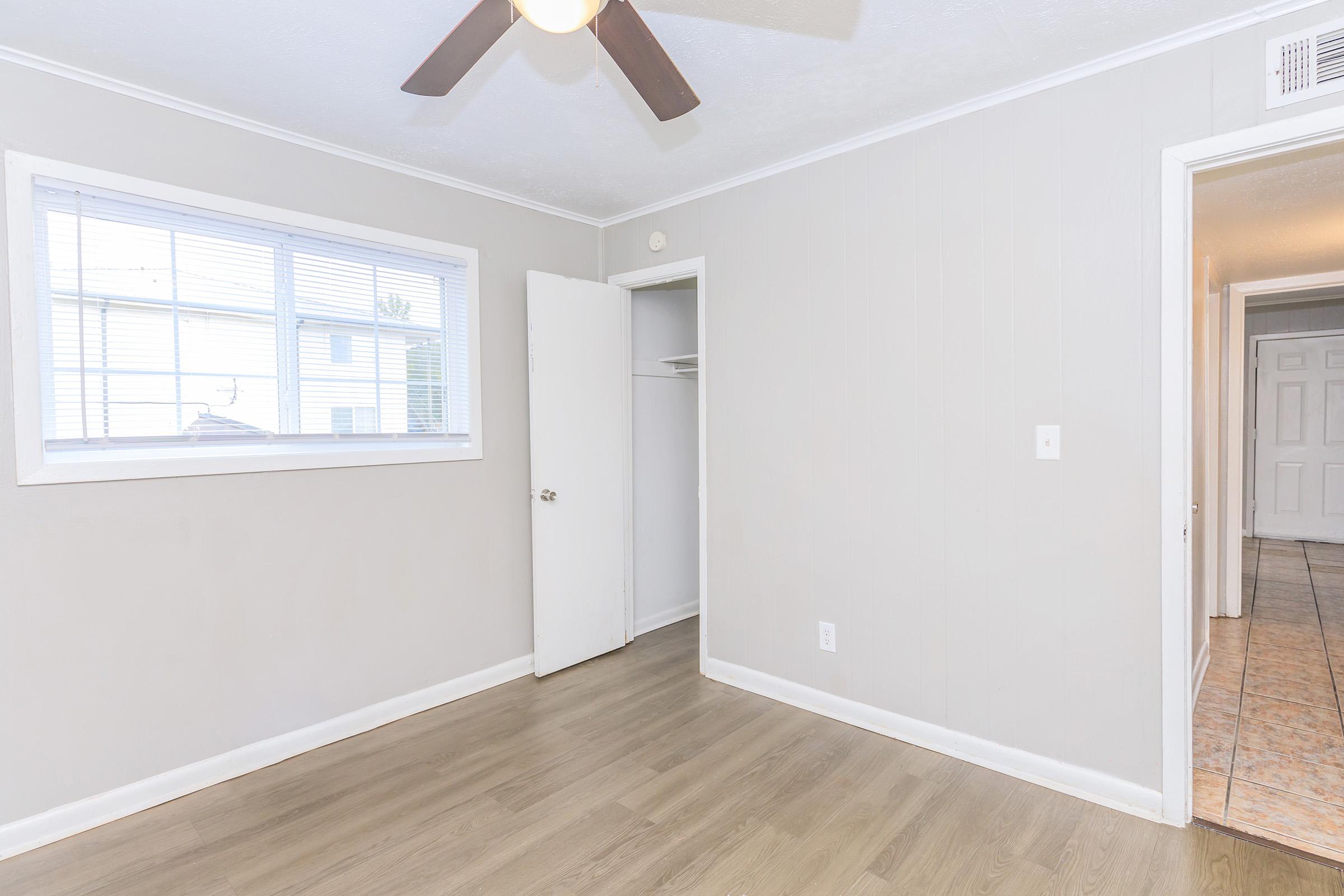 Empty room featuring light gray walls, a ceiling fan, and a window with blinds allowing natural light. A closet door is partially open, and there's a doorway leading to another room. The flooring is light-colored, possibly laminate or hardwood, contributing to a bright and spacious feel.