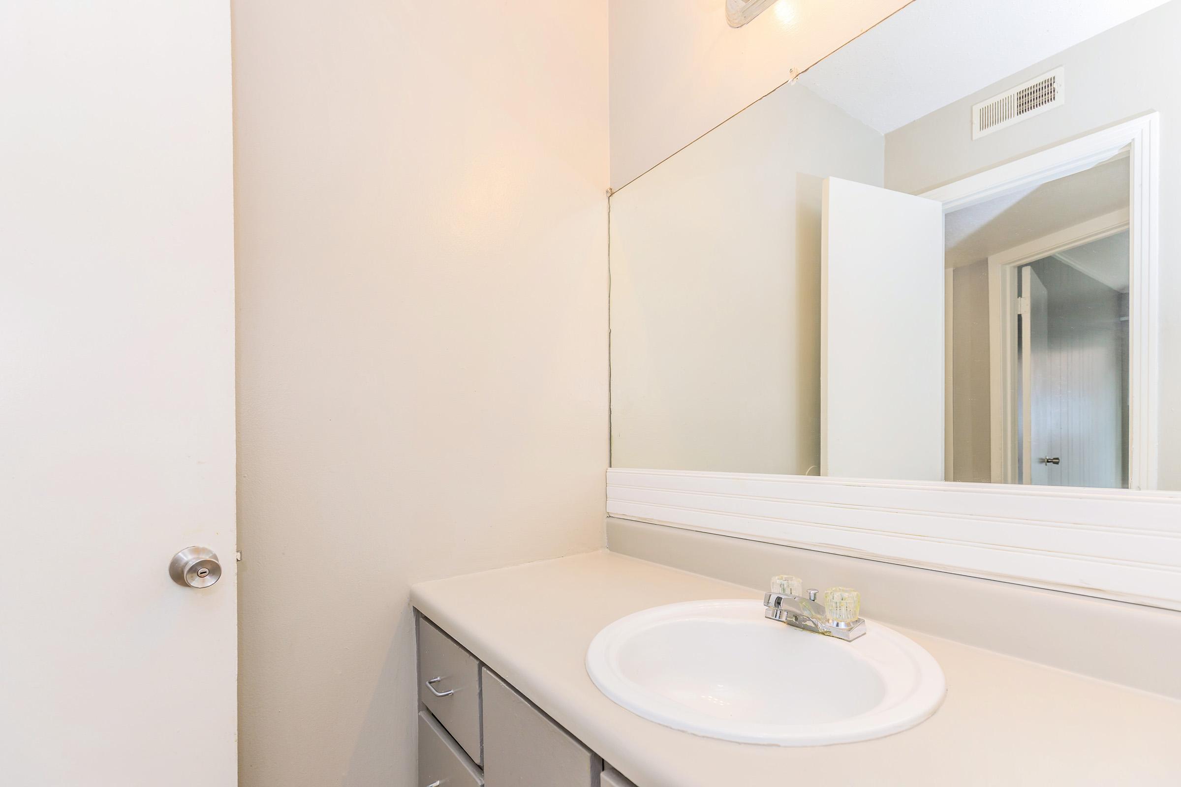 A clean bathroom with a light-colored countertop and a round sink. There is a mirror above the sink reflecting the room. The wall is painted in a soft hue, and a door is visible on the left leading to another area. Natural light brightens the space.