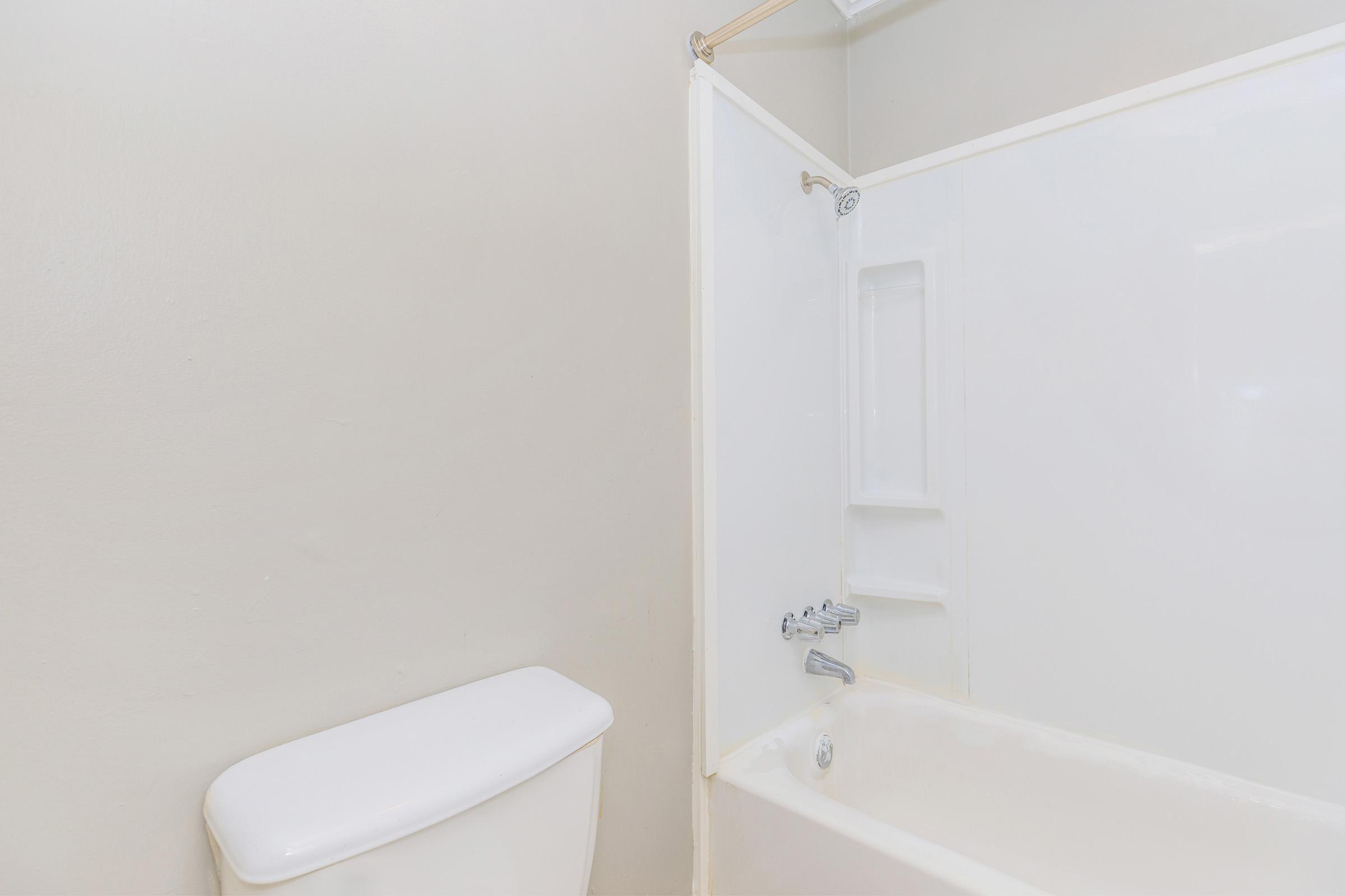A clean and simple bathroom featuring a white bathtub with a shower, a white toilet, and light-colored walls. The bathtub has a chrome faucet and a built-in shelf for storage. The overall design is minimalistic and spacious.